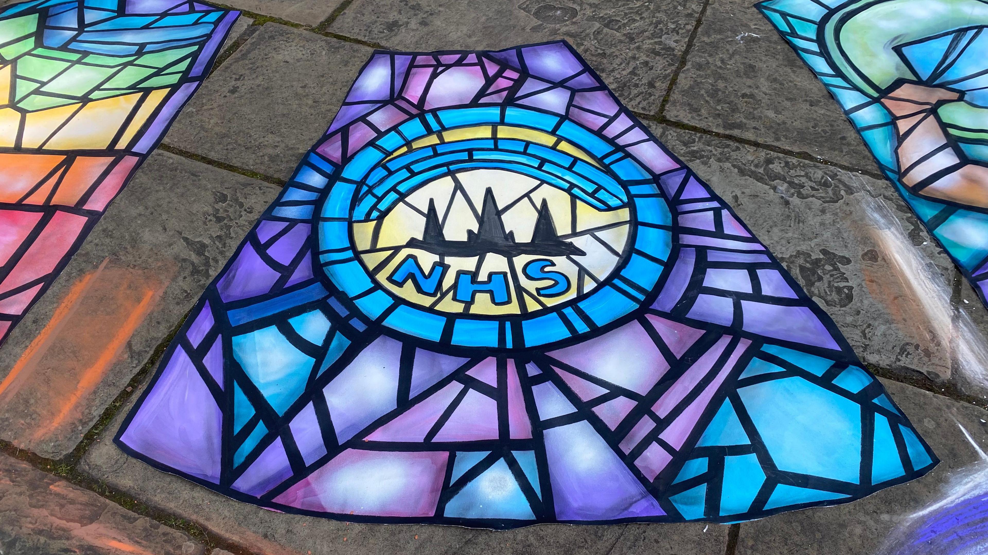 A section of an artwork created on the floor, resembling the pane of a stained glass window. It  contains the letters NHS in blue and a silhouetted skyline.