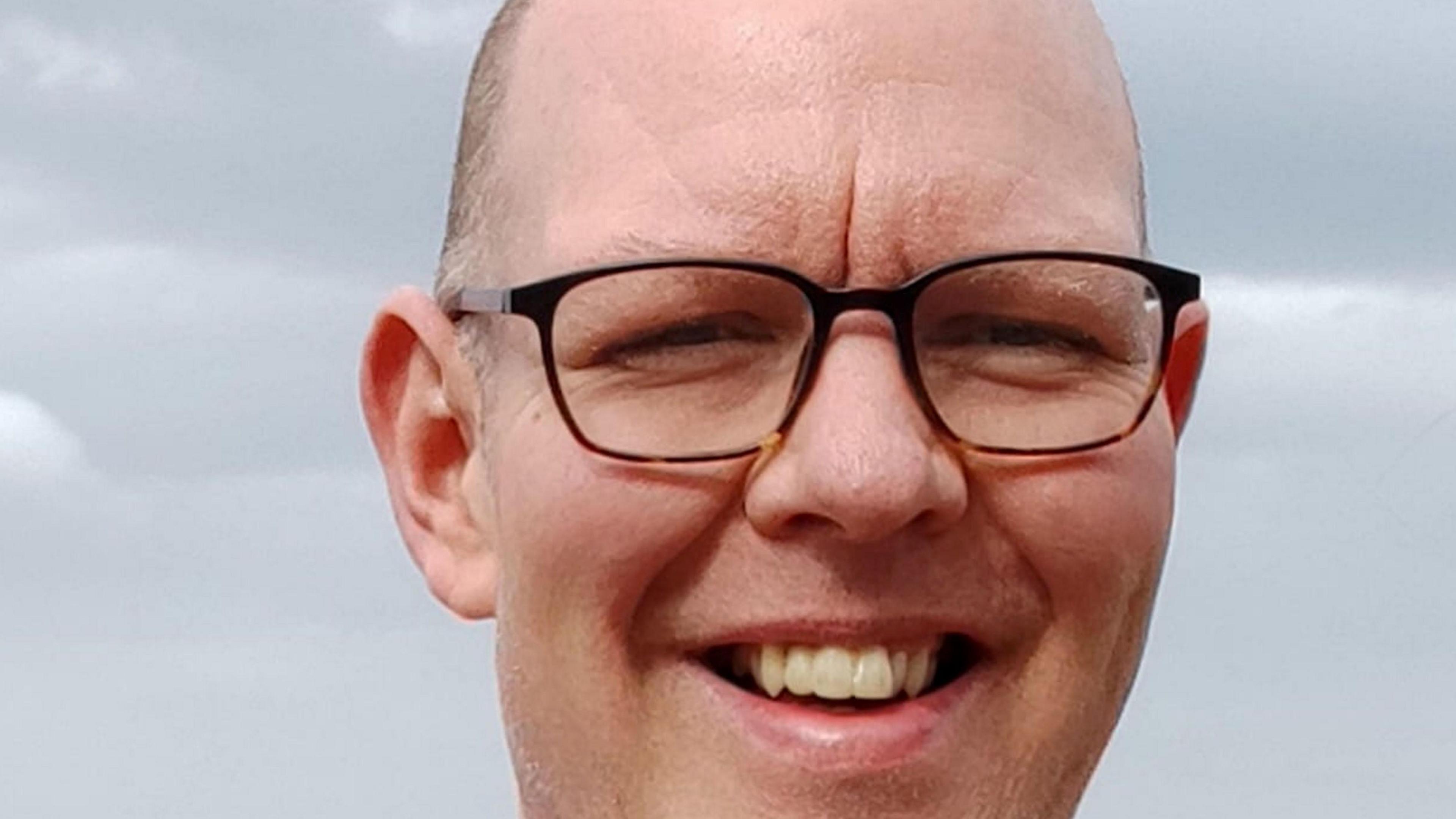 A bespectacled man smiles widely at the camera. He appears to be standing outside, with a grey sky in the background.