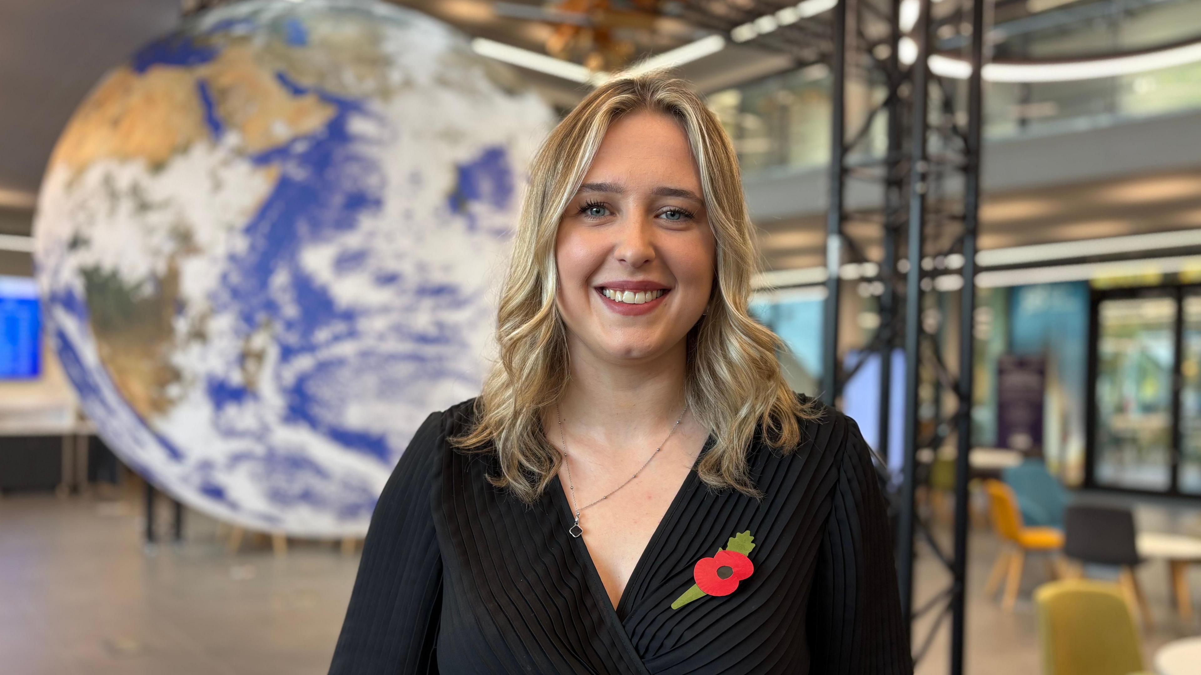 Research Associate Abigail Waring has shoulder-length blonde hair. She is wearing a black top and a poppy. A model of Planet Earth is behind her.