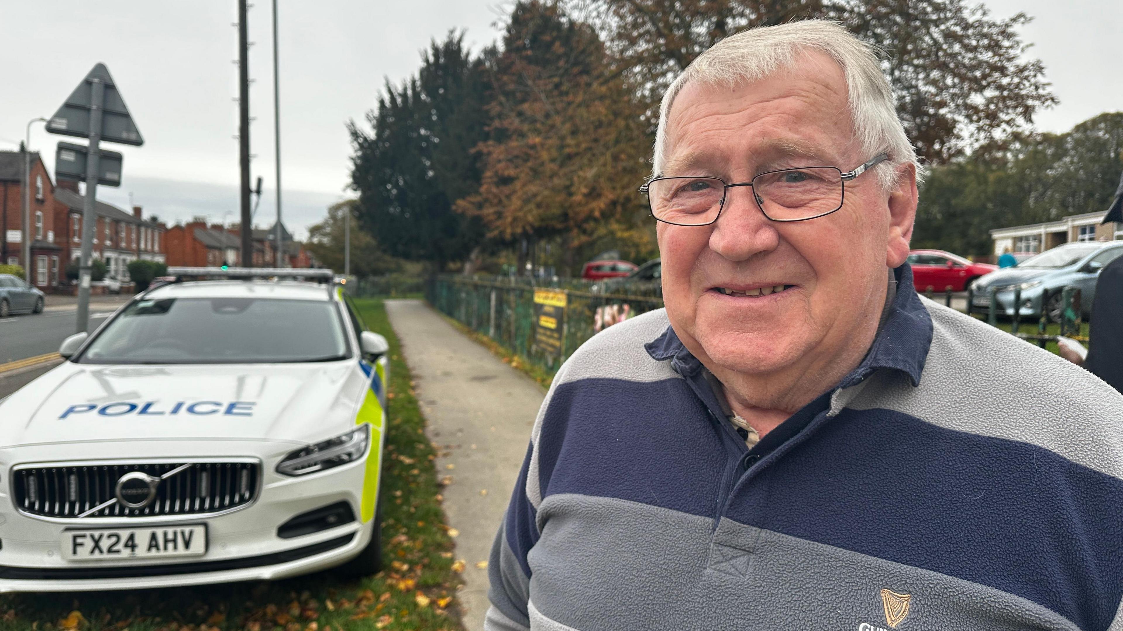 A man wearing a blue striped top and glasses is looking into the camera. There is a police car in the background.