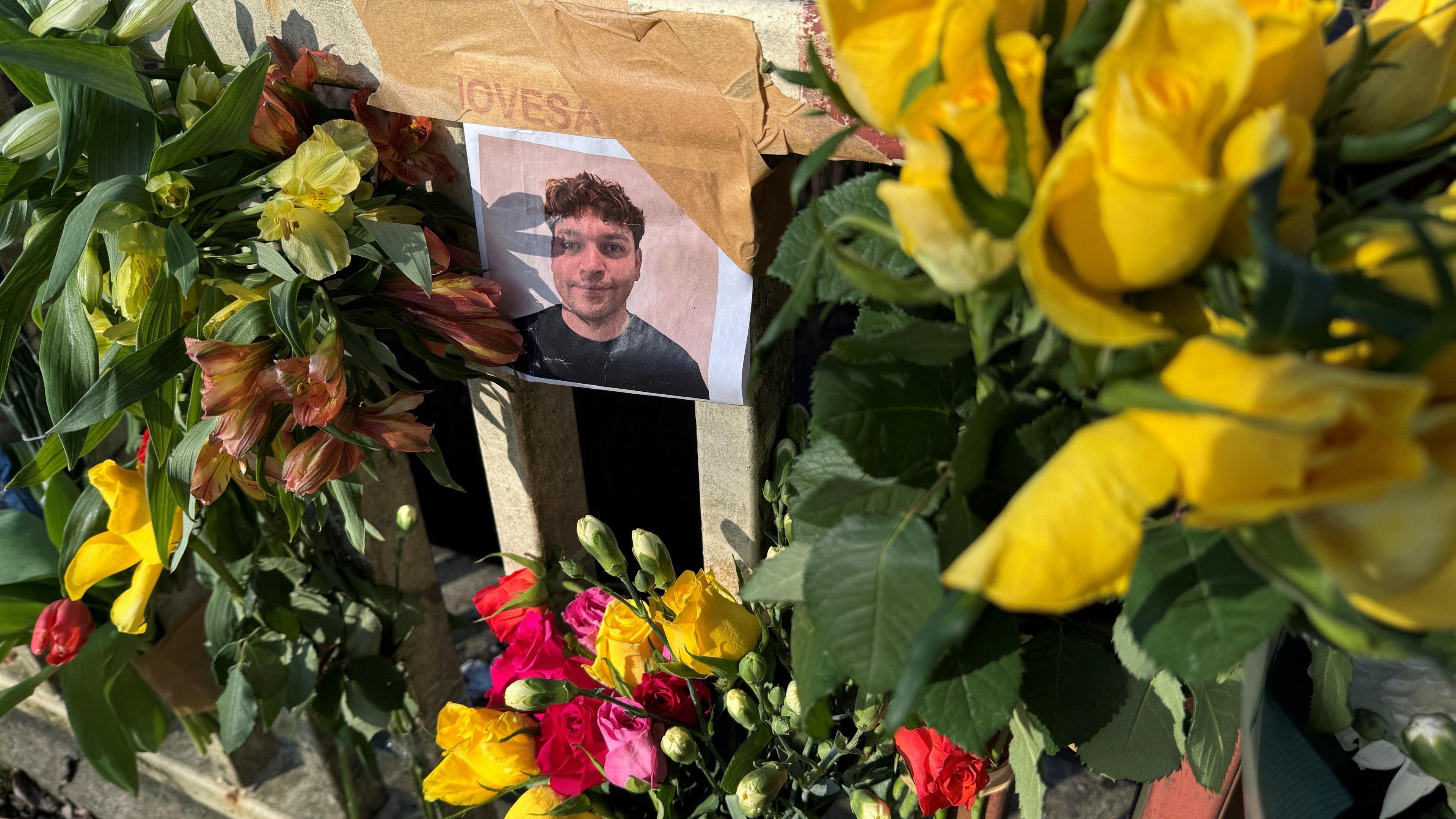 A picture of Mr Piovesana taped to some metal railings beside a river. The image is surrounded by bouquets of colourful roses pink, yellow and red roses.