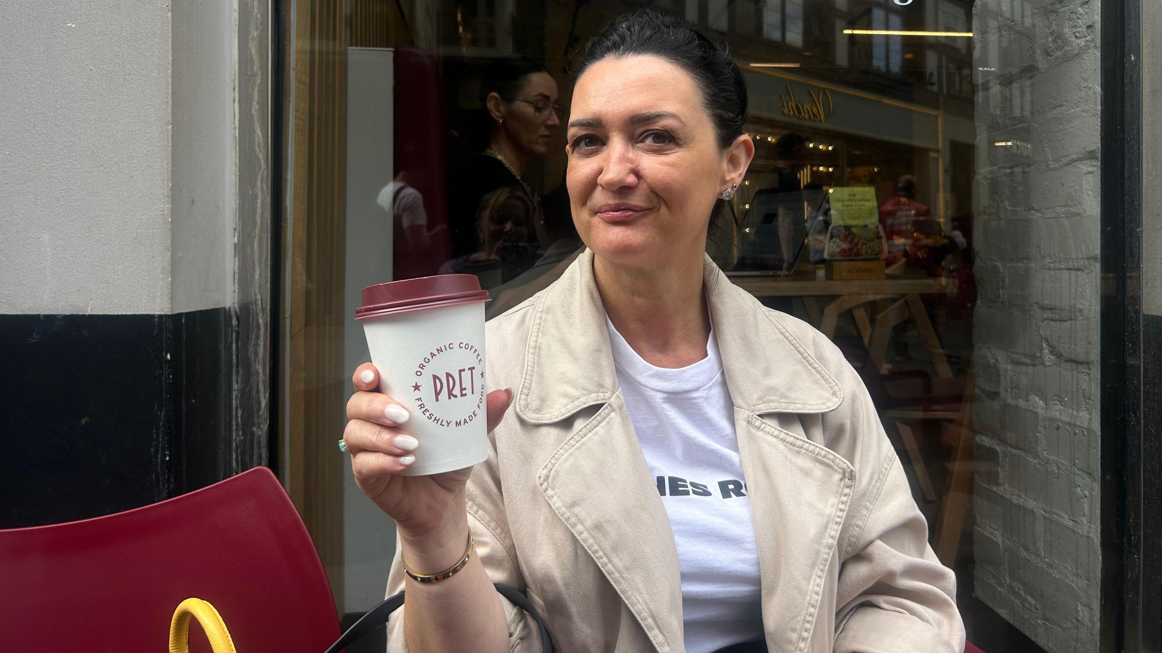 Mimoza Emsa with dark brown hair in a ponytail wearing a beige jacket over a white t shirt sitting outside Pret holding a takeaway coffee cup