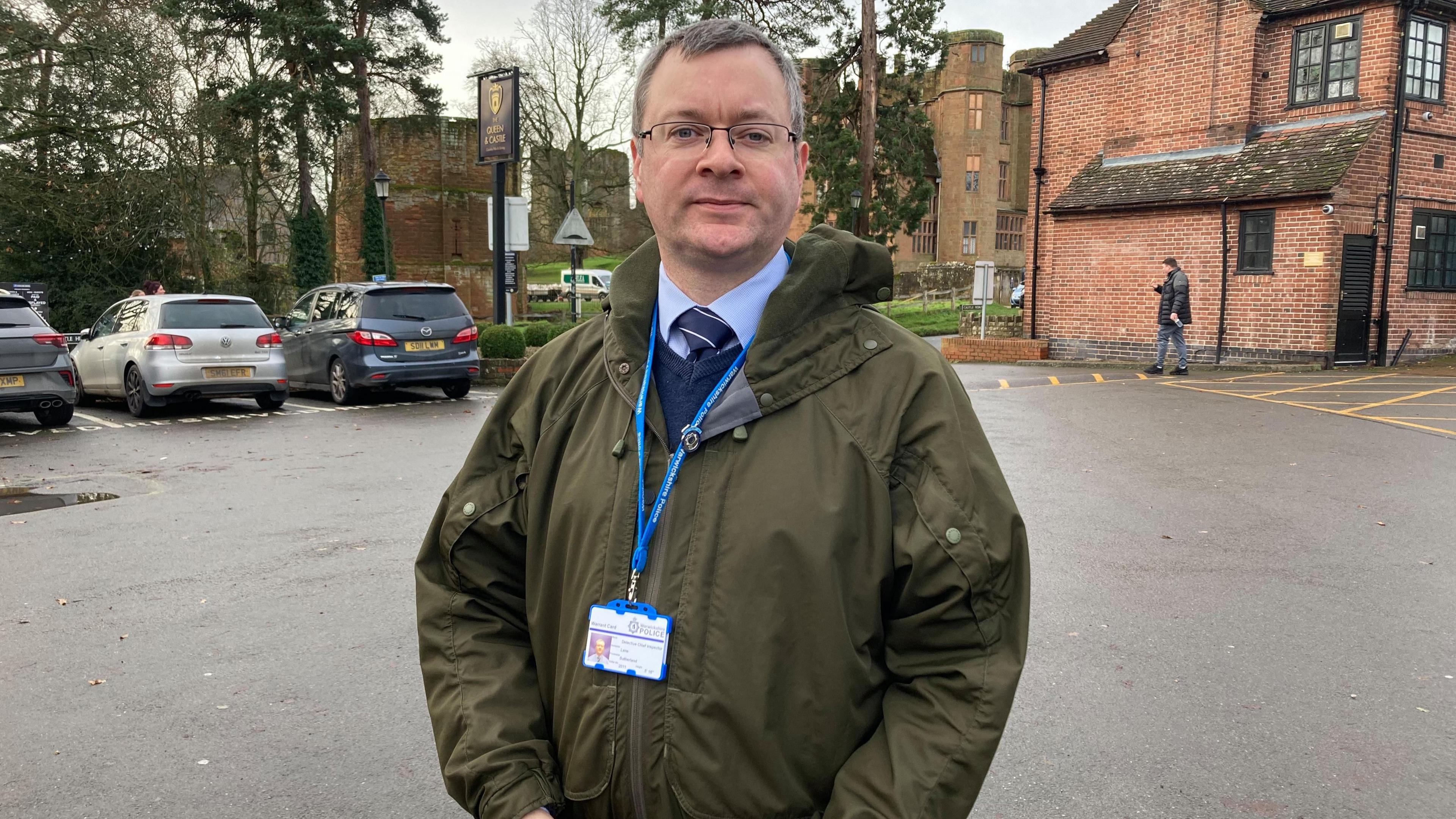 A man with grey hair and glasses stands in a car park looking at the camera. He is wearing a blue shirt, blue tie and blue pullover and a green coat. He is wearing an official-looking lanyard.