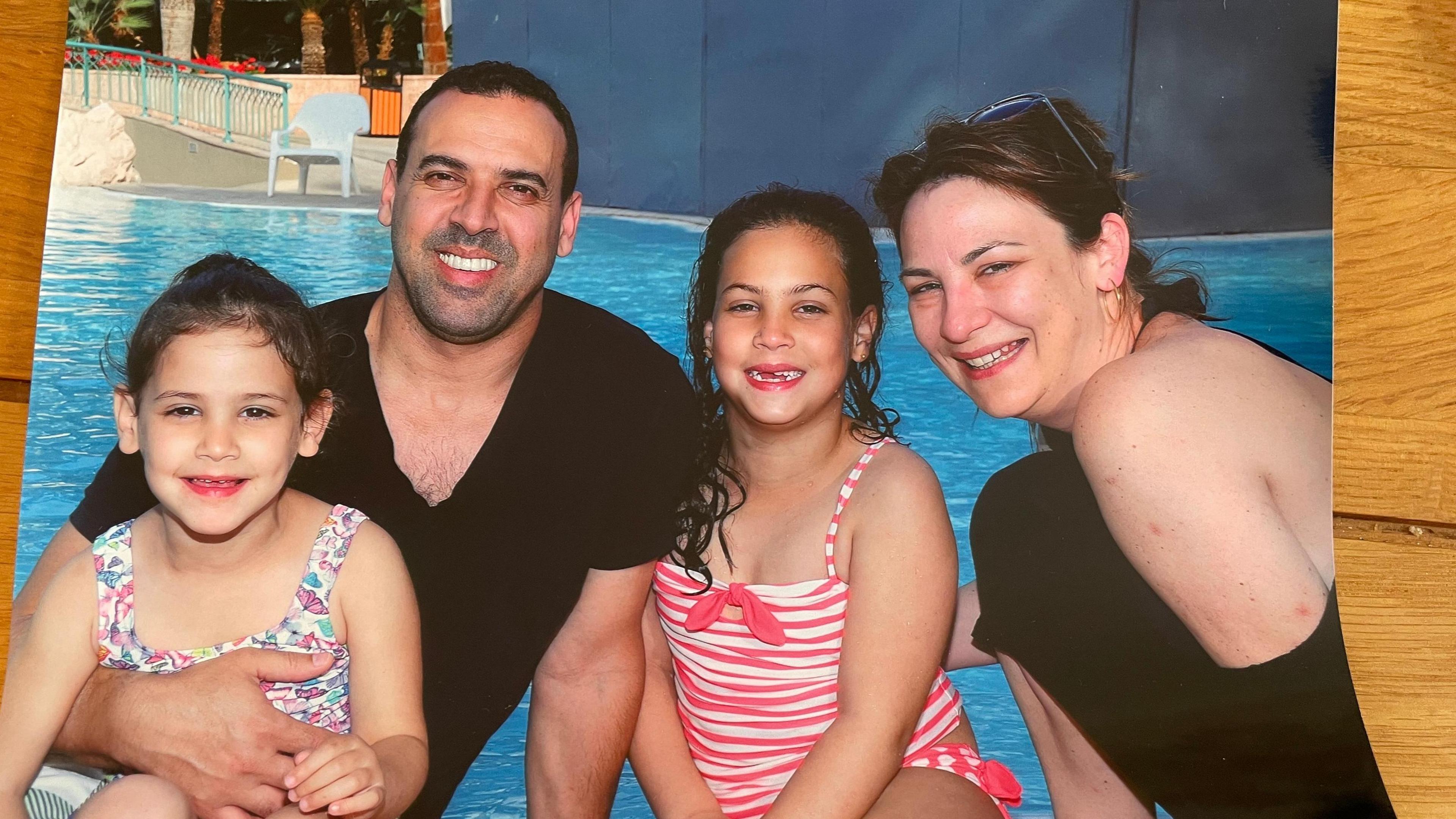 A family of four sit in front of a swimming pool. The man holds a young girl on his lap, an older girl sit next to him, with his wife on the right of the picture leaning in. 