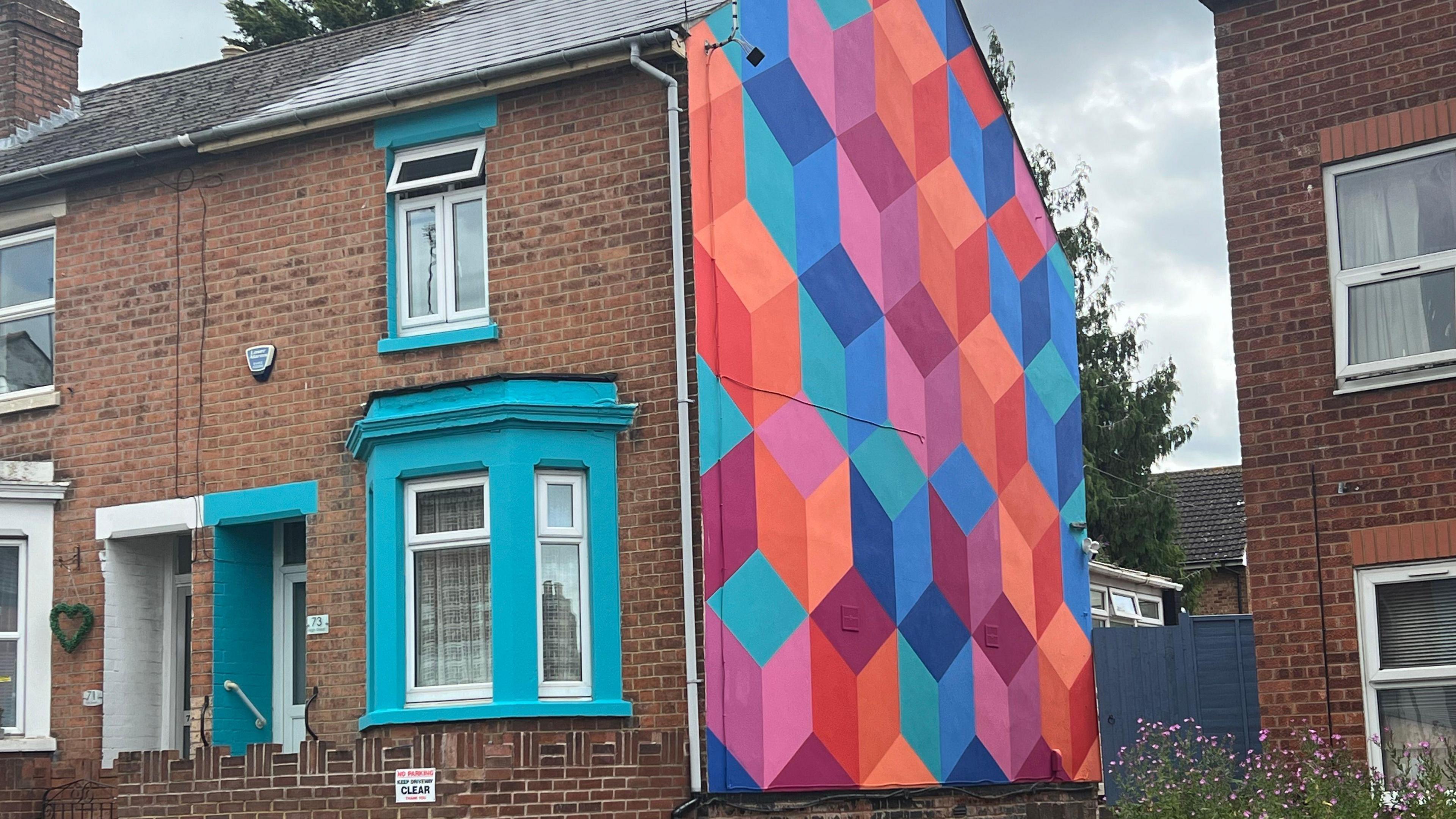 A red brick house with a colourful mural on the side. It is patterned with cubes, painted in red, orange, pink, purple and blue.