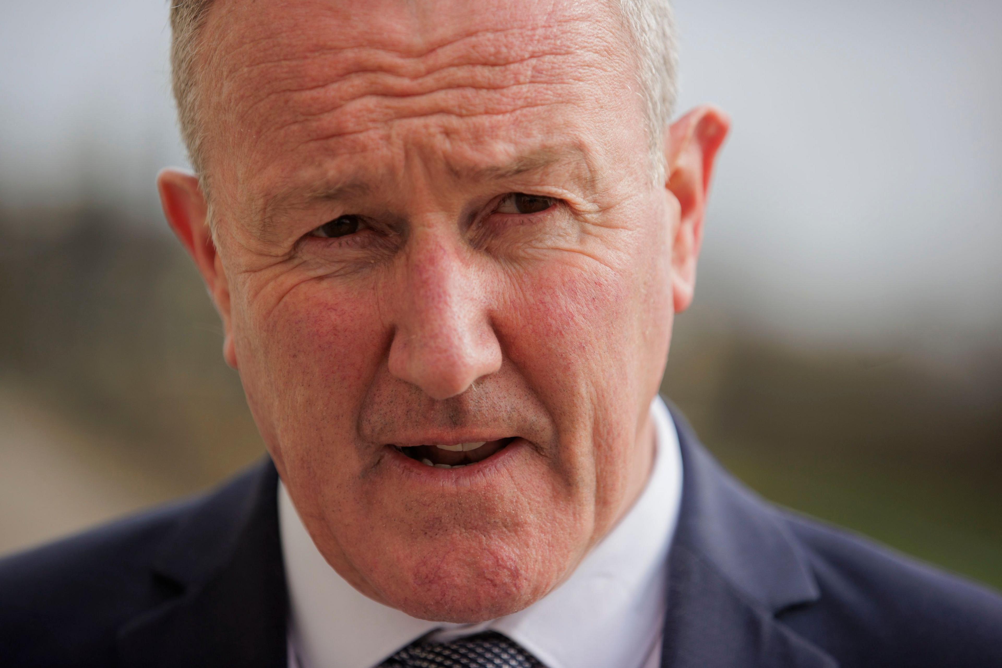 Northern Ireland Economy Minister Conor Murphy at Parliament Buildings. He's wearing a dark suit with a white shirt and his tie is black and white. He's standing outside, but the background is blurred. The camera has captured him while speaking, so his mouth is partially open.