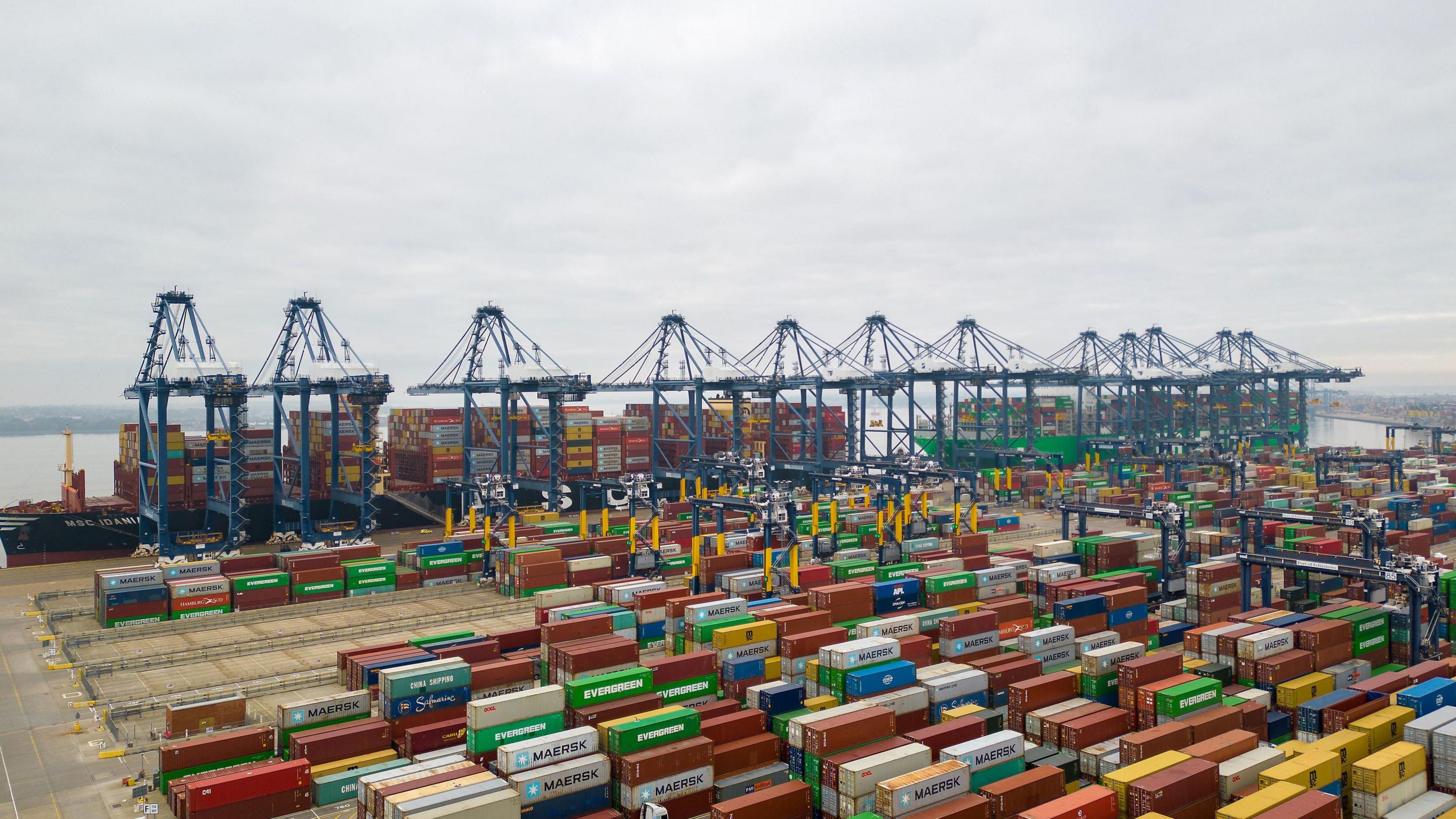 A large number of shipping containers sit in front of loaded boats and tall blue machinery. The sea is visible in the background. 