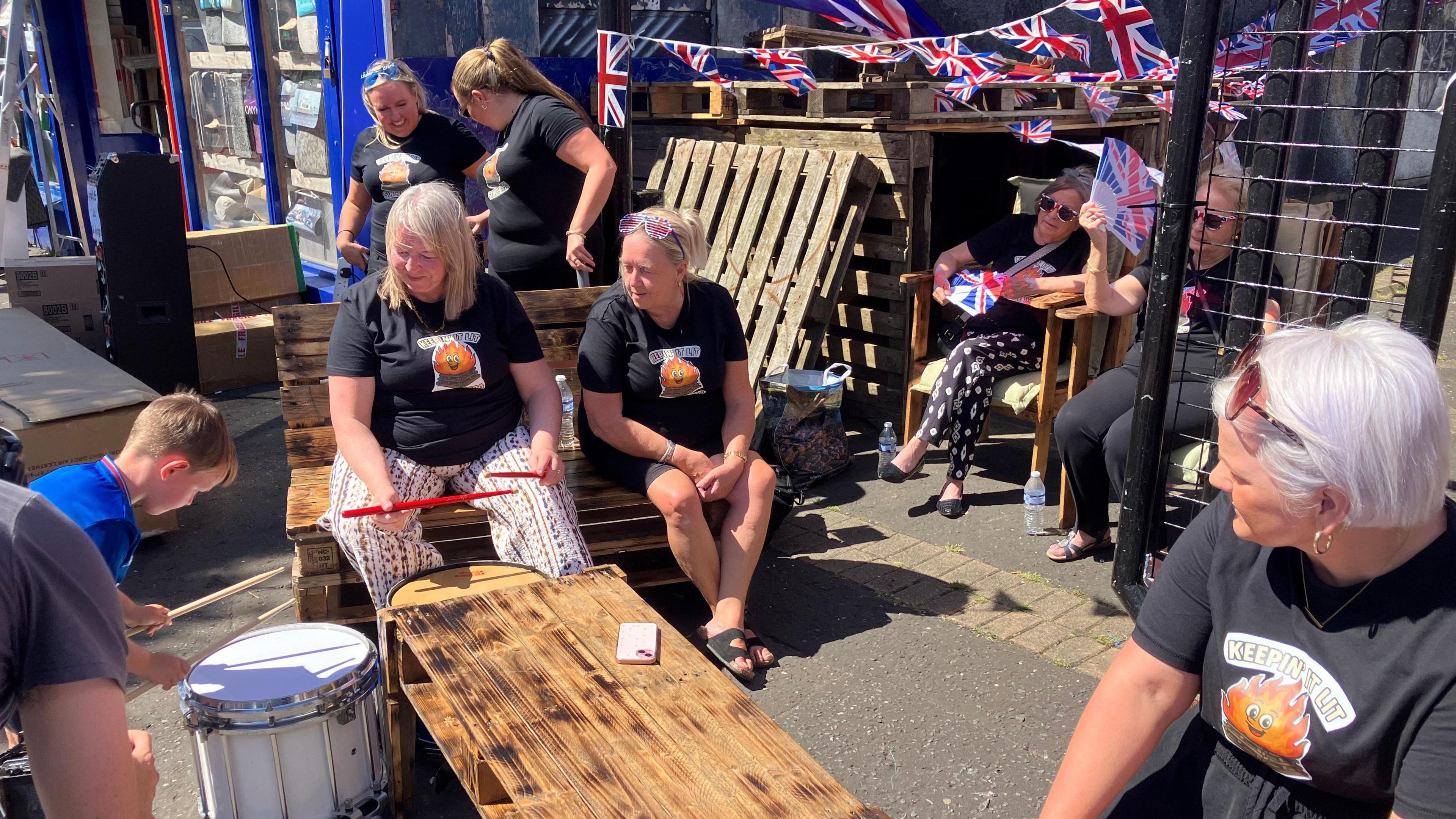 A group of women are sitting outside and watching a young boy play a drum. They are all wearing black t-shirts which feature an image of a smiling bonfire and the words 'Keepin It Lit'. 