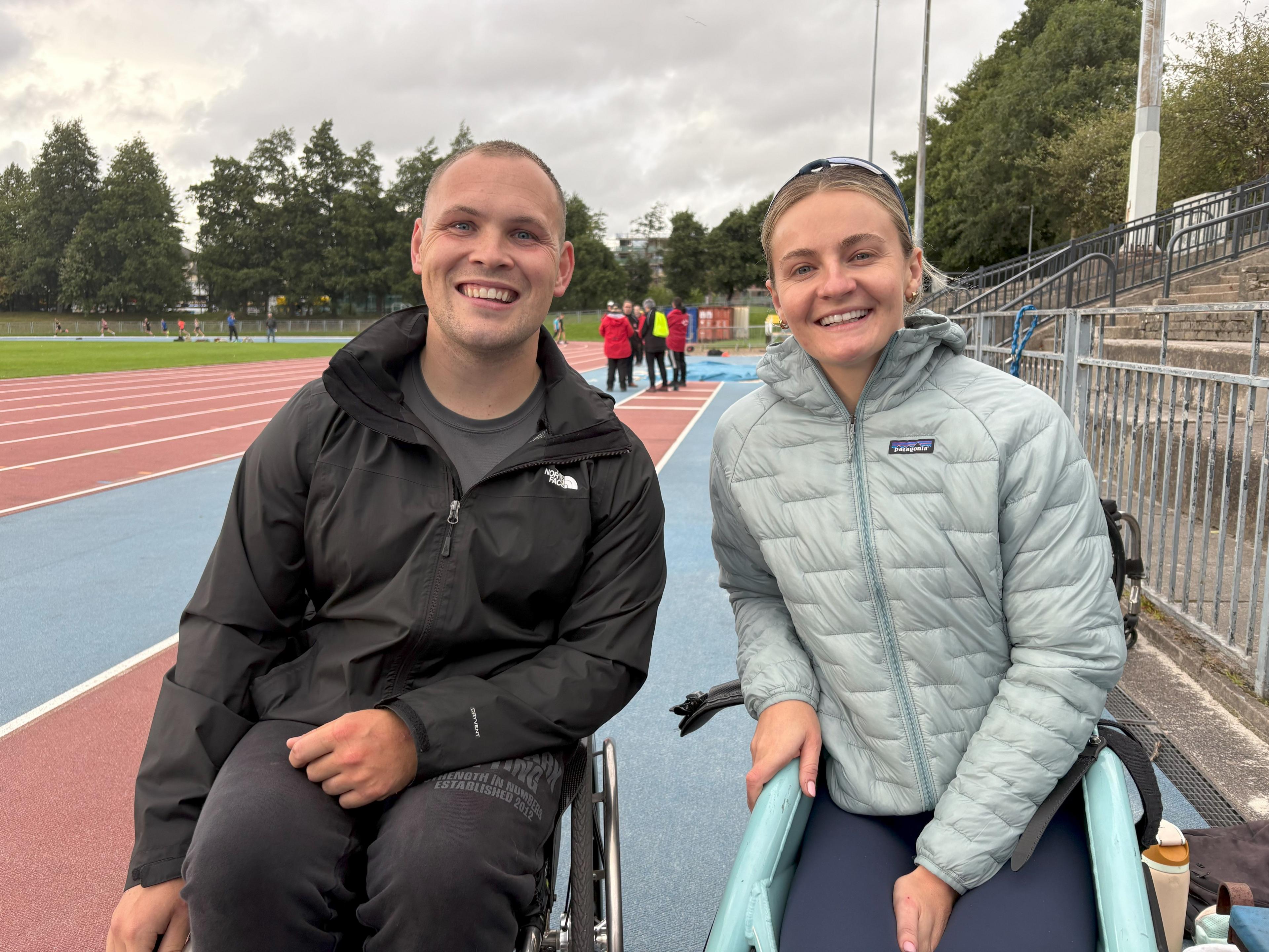 Image of wheelchair racers Ben Rowlings and Melanie Wood at their running track. The pair are smiling and wearing outdoor jackets