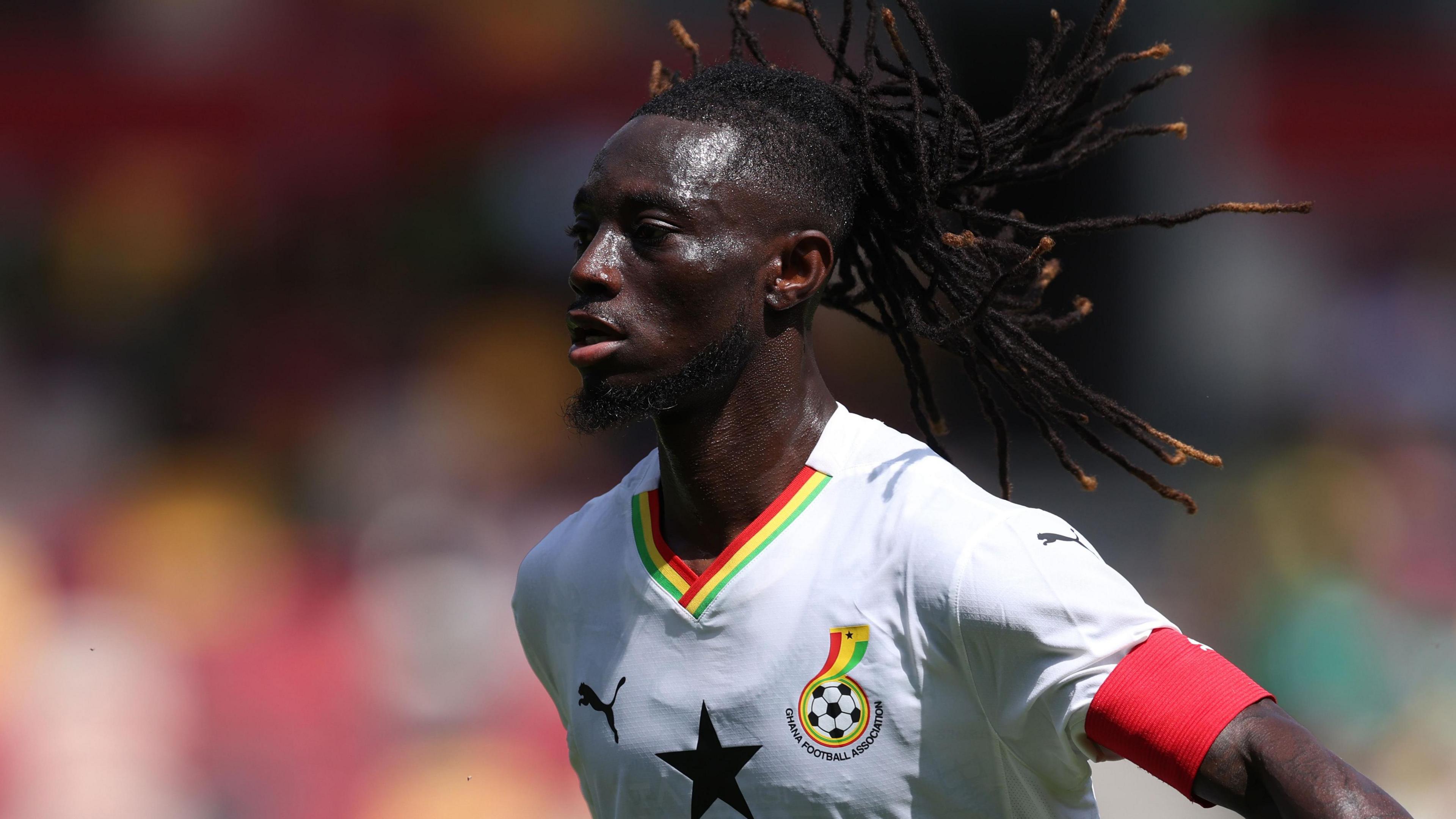 Gideon Mensah, wearing a red armband on his left arm and a white Ghana shirt with a black star on the front, is seen from chest up during a football match. His black braids with orange tips are seen flowing behind him as he moves