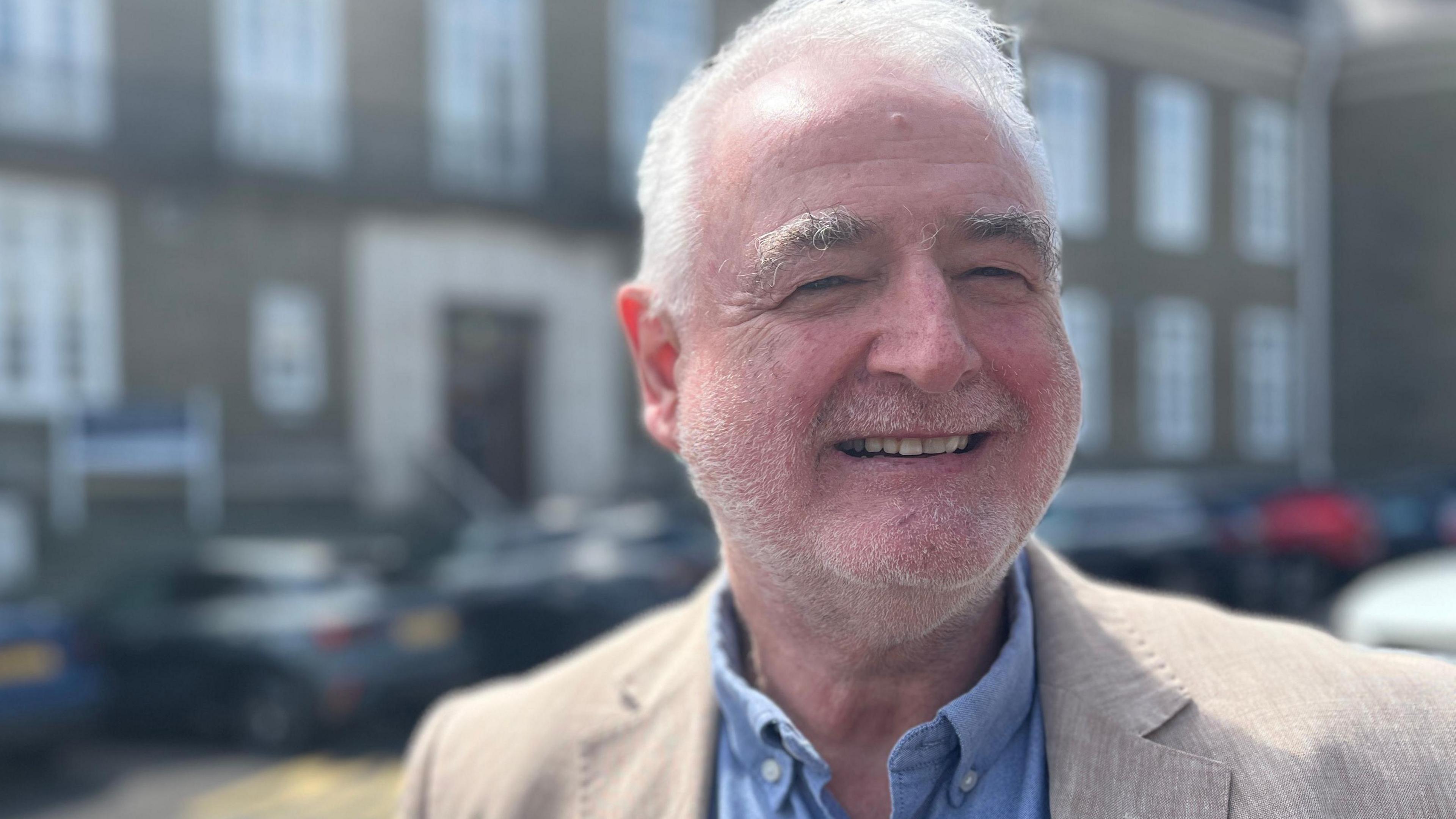 Owen Jenkins in a beige tweed jacket and blue shirt smiles looking slightly off camera. He has short grey hair and grey stubble.