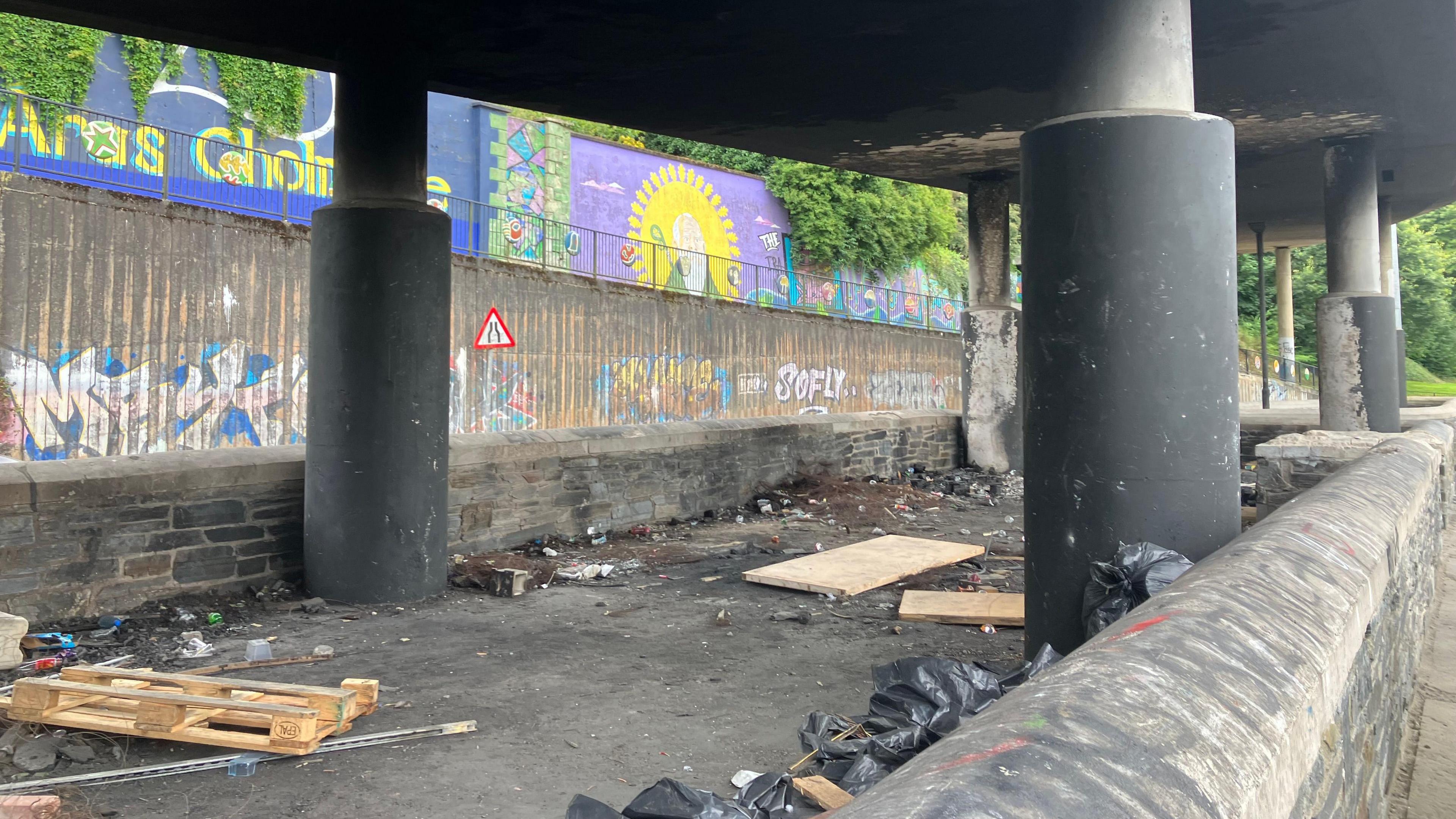 Concrete columns hold up a flyover type road. a number of wooden pallets and other debris can be seen in the space under the road