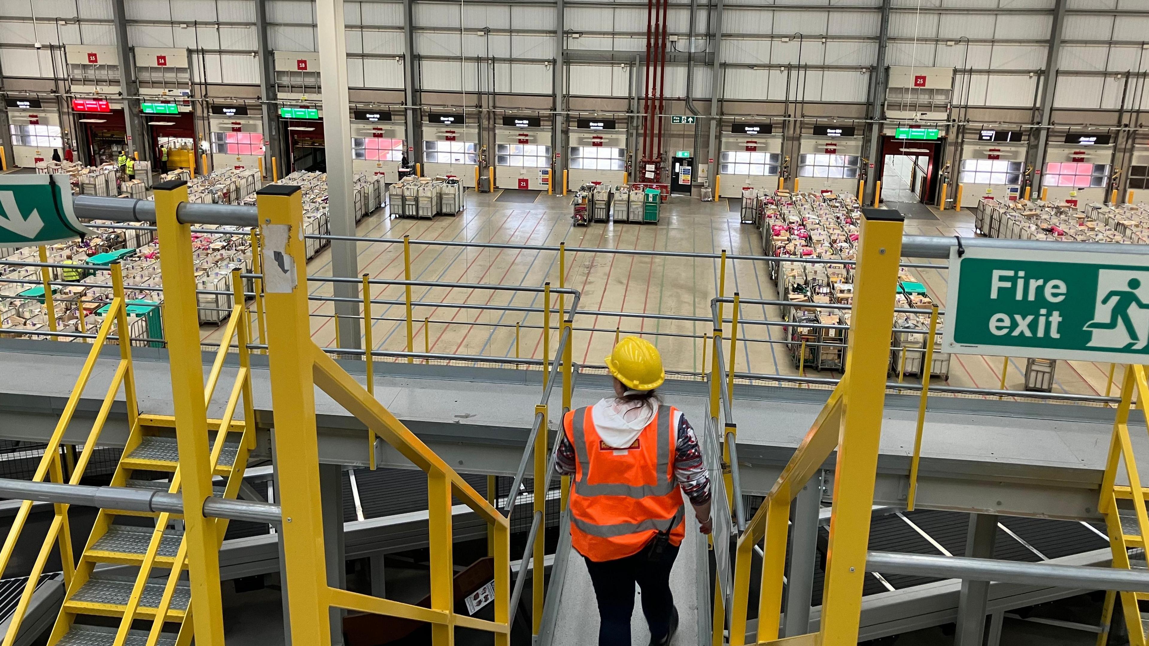 A person in a high-viz jacket and a hard hat stands on a gantry looking across a warehouse. There are hundreds of parcels on a level below them.