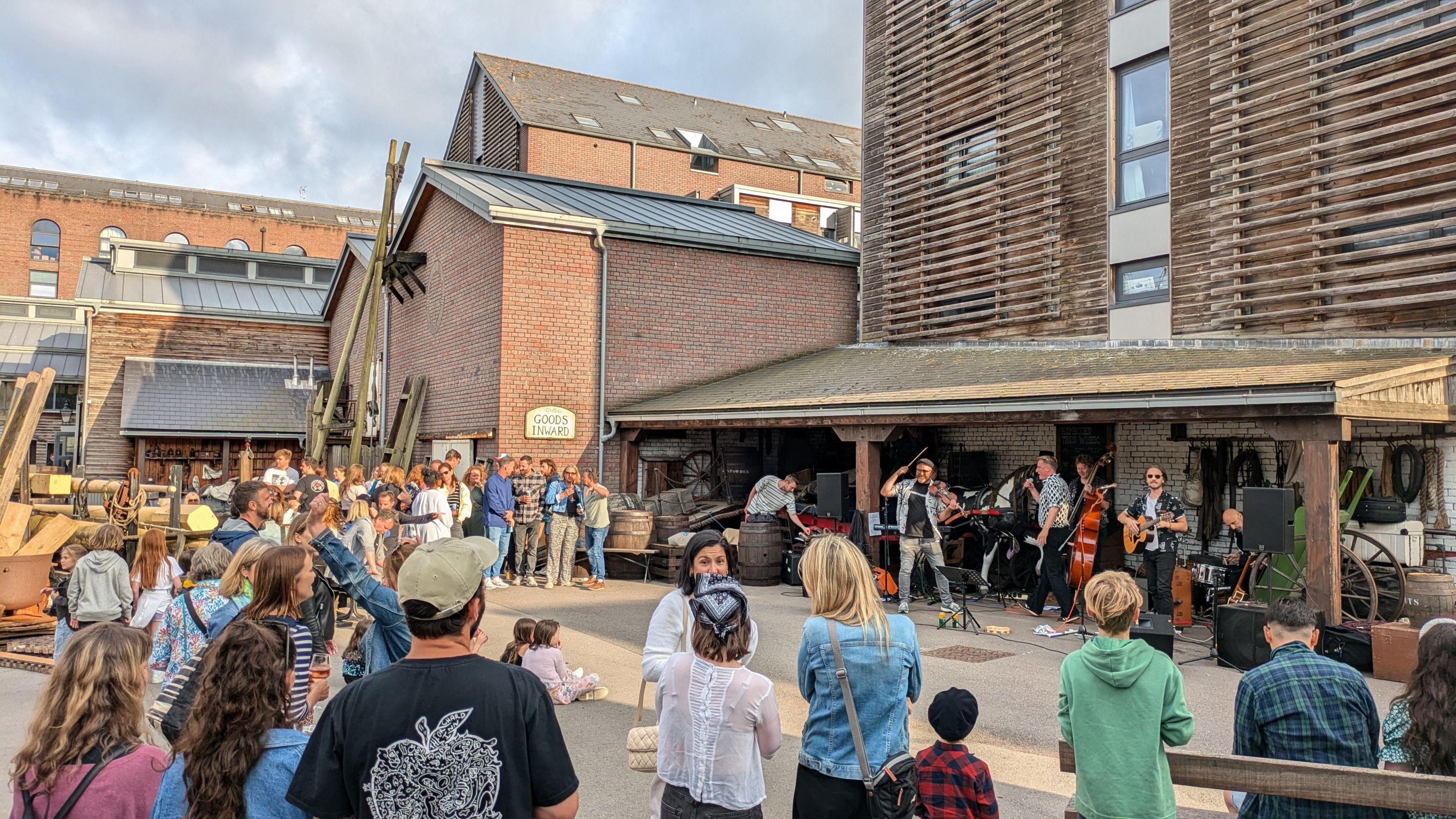 A group of people stand around in a circle watching musicians play in the dockyard at the ss Great Britain in Bristol