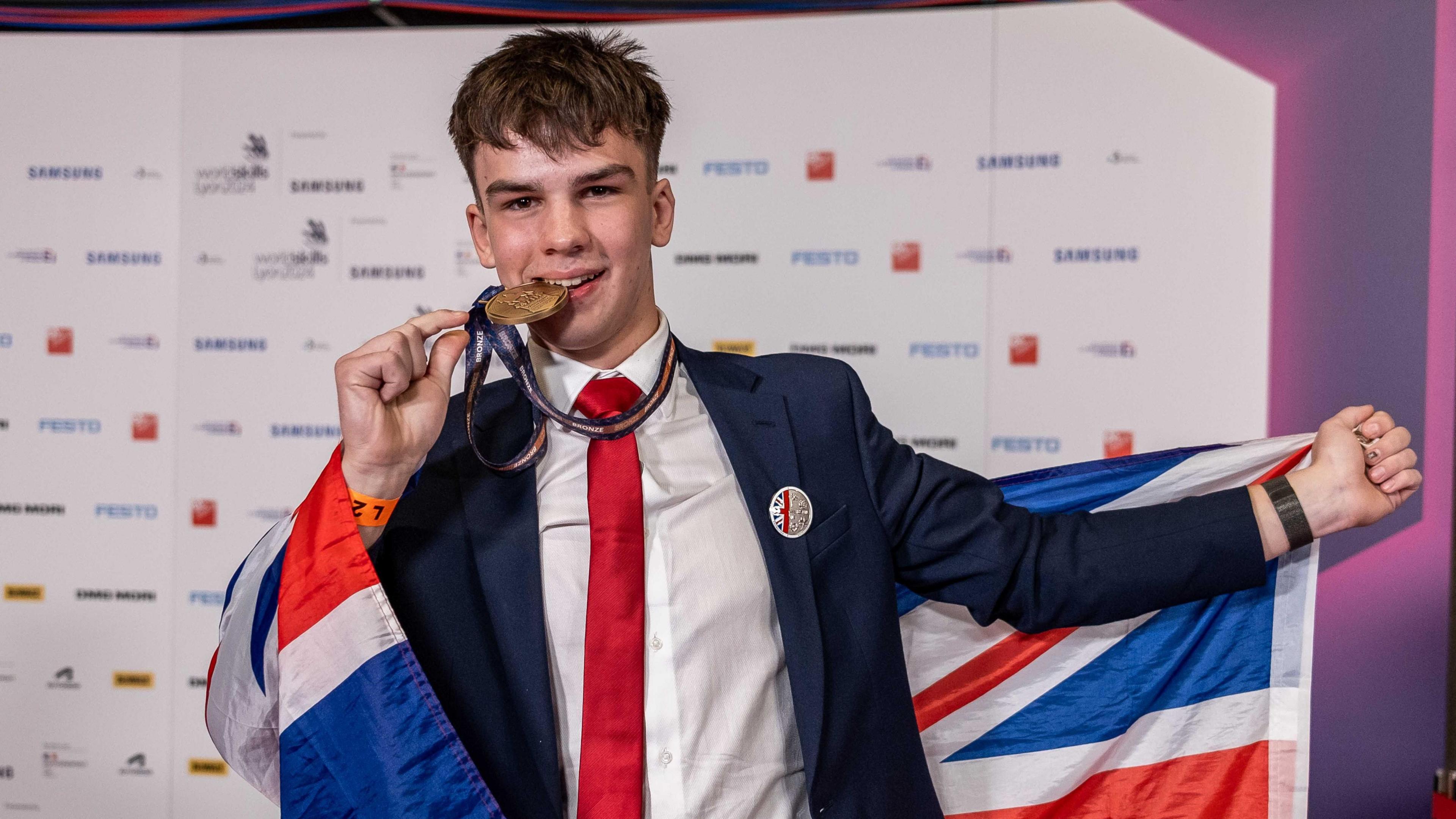 A man dressed in a navy suit with a white shirt and red tie. He is pictured holding and pretending to bite a bronze medal whilst holding a Union Jack flag.