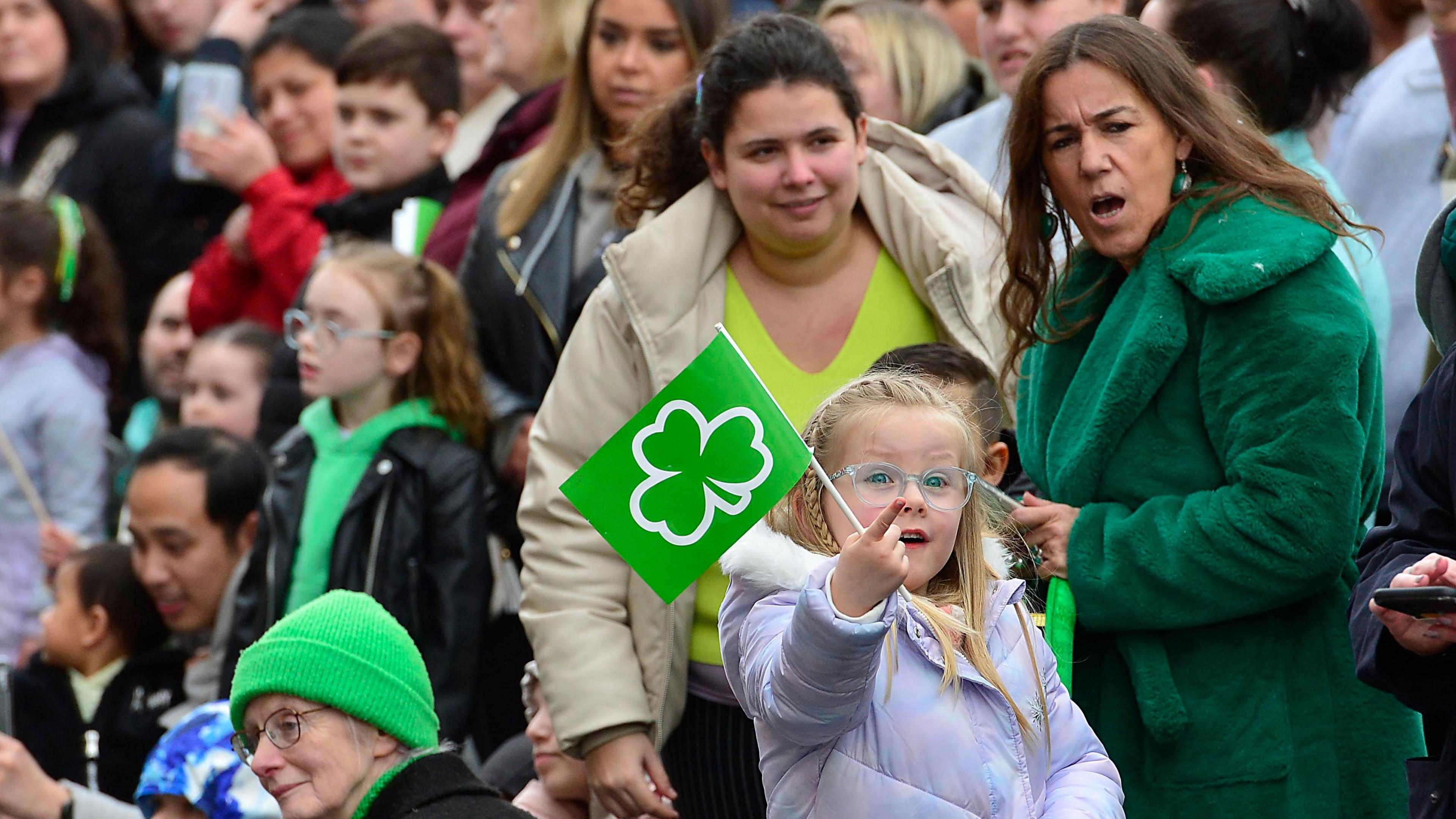 excited girl holding shamrock flag points at something coming her way while everyone around her looks in the opposite direction