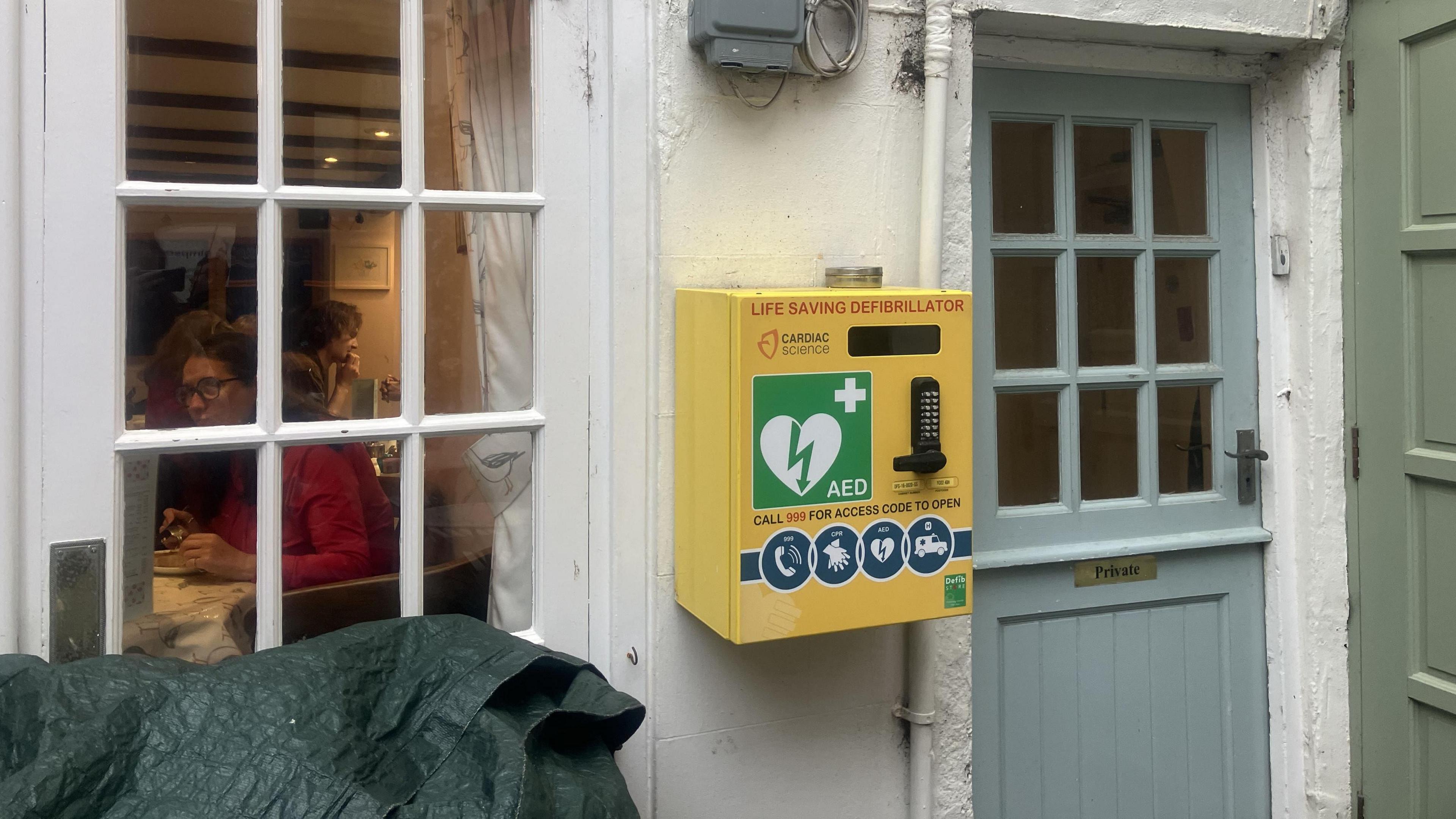 On a white wall is a defibrillator. It is yellow with a pin pad on the front. In the background people are eating in the café. 