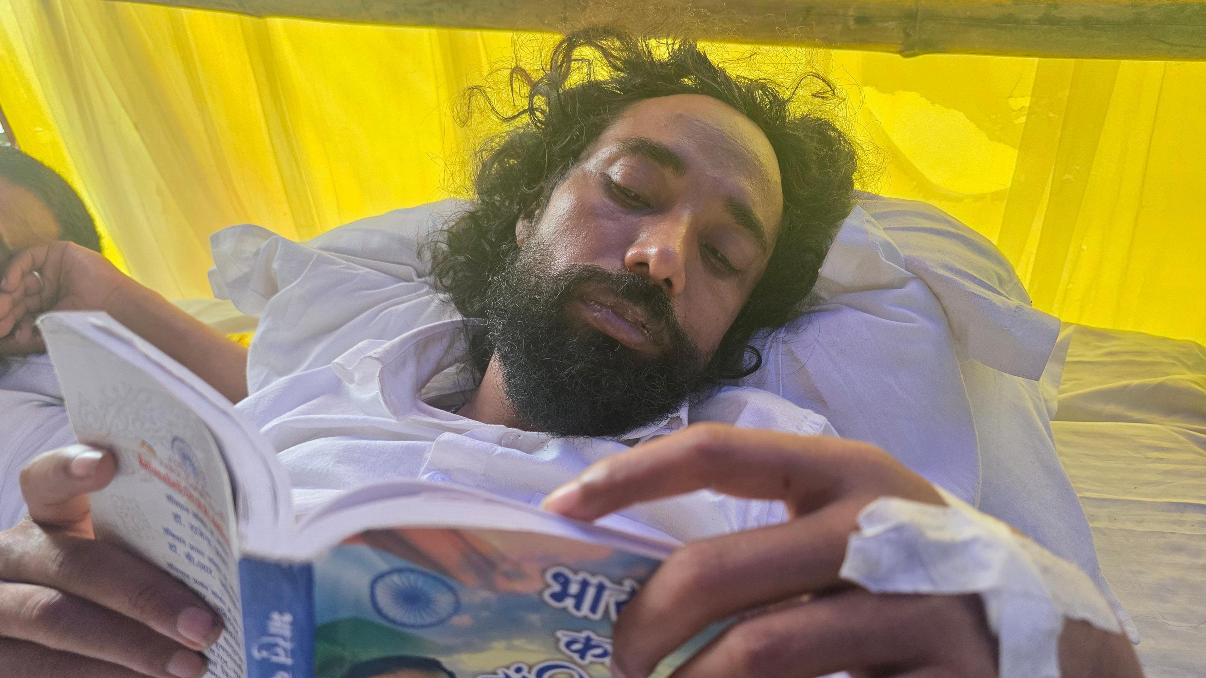 Devendra Nath Mahto laying on a mattress wearing white clothes and reading a book 