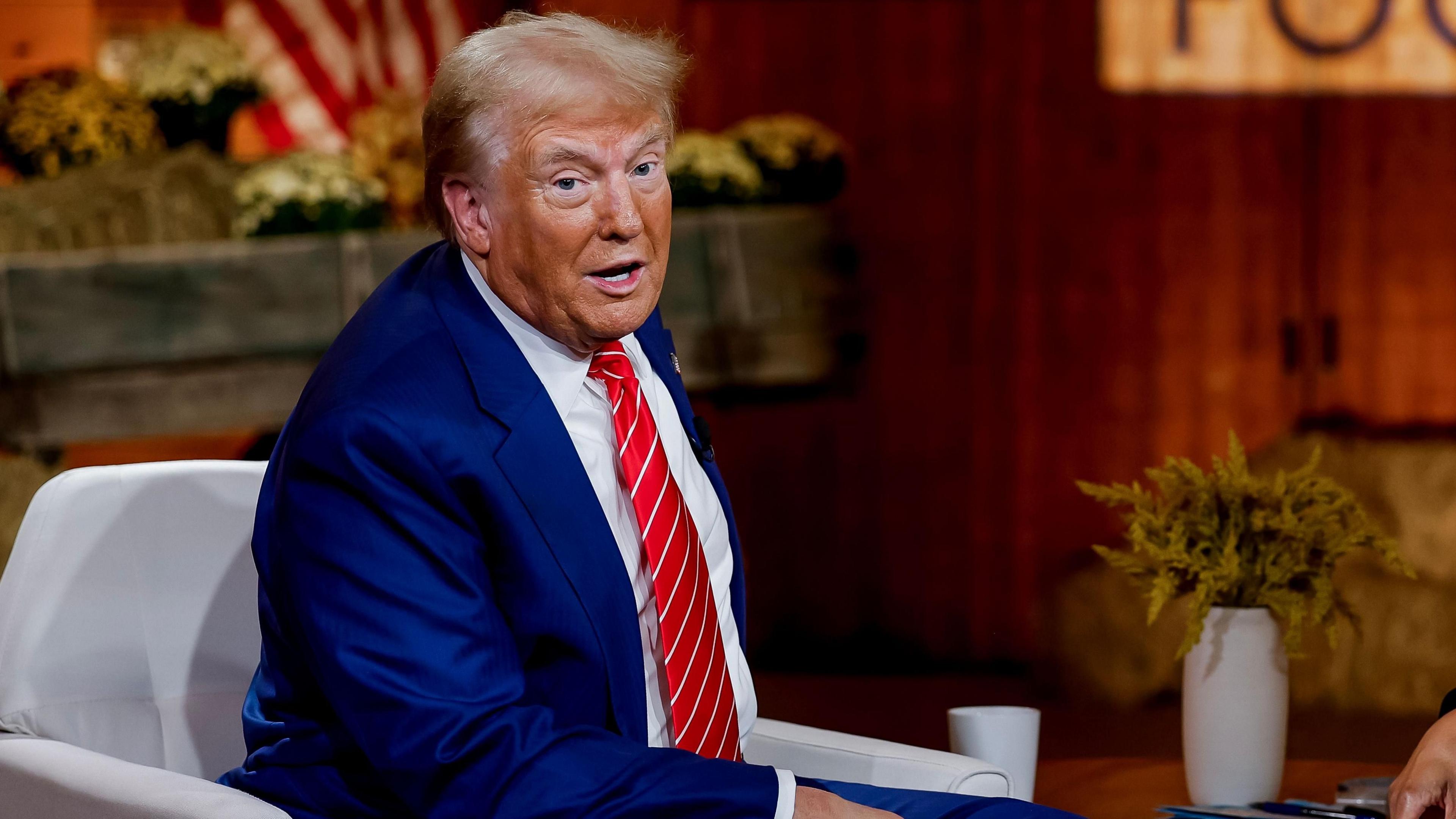 Trump in blue suit with red tie seated in front of American flag and plants