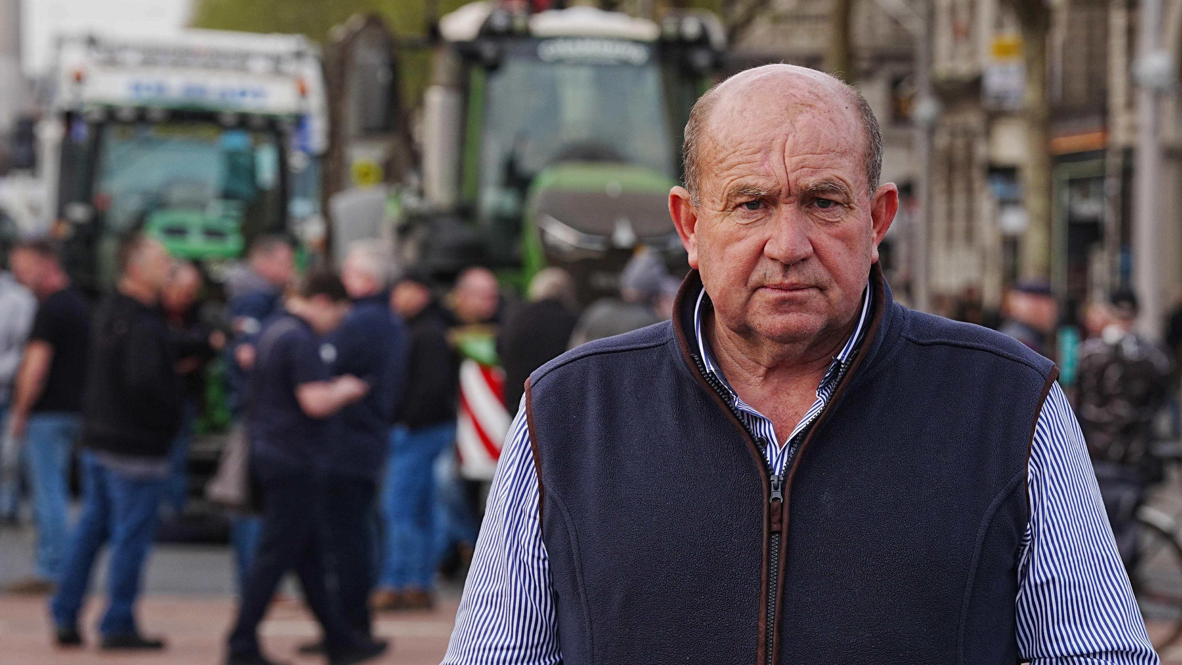 A man looks at the camera. He has a serious look on his face. He is wearing a blue and white shirt and a dark-blue gilet jacket. Behind him are two green tractors parked in an urban area. 