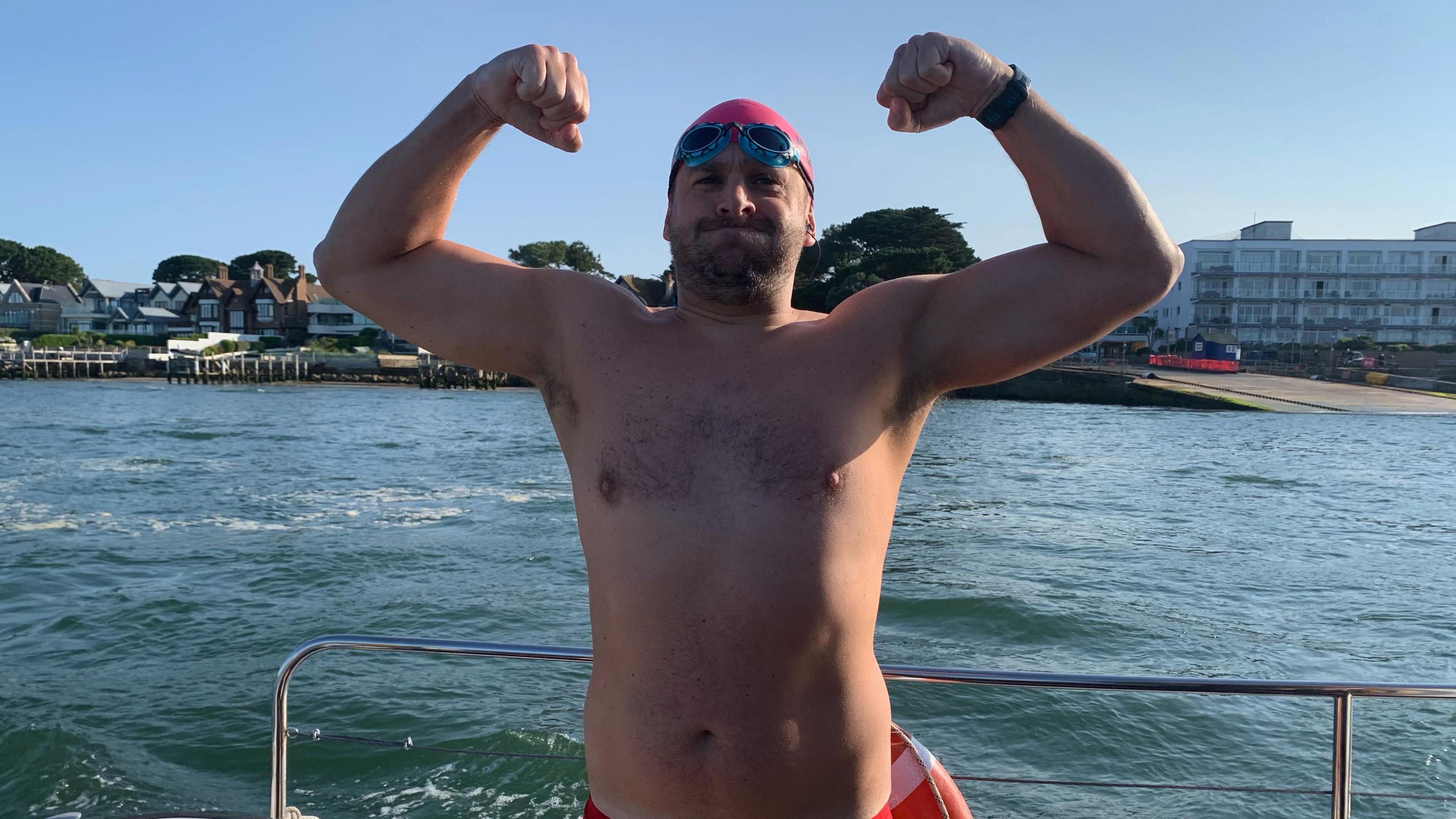 Alexander David on a boat lifting both his arms to flex. He is wearing a swim cap and goggles. The sea and coastline are behind him, with a number of houses and a hotel visible.