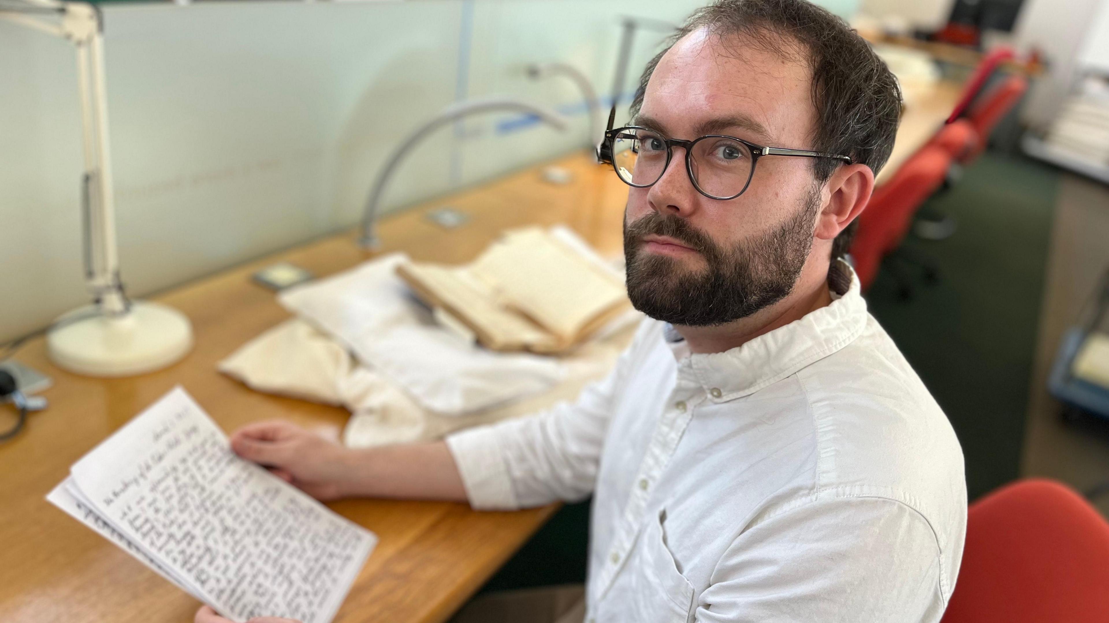 A bearded man with a white button down collar shirt at a desk holding a sheaf of paper.