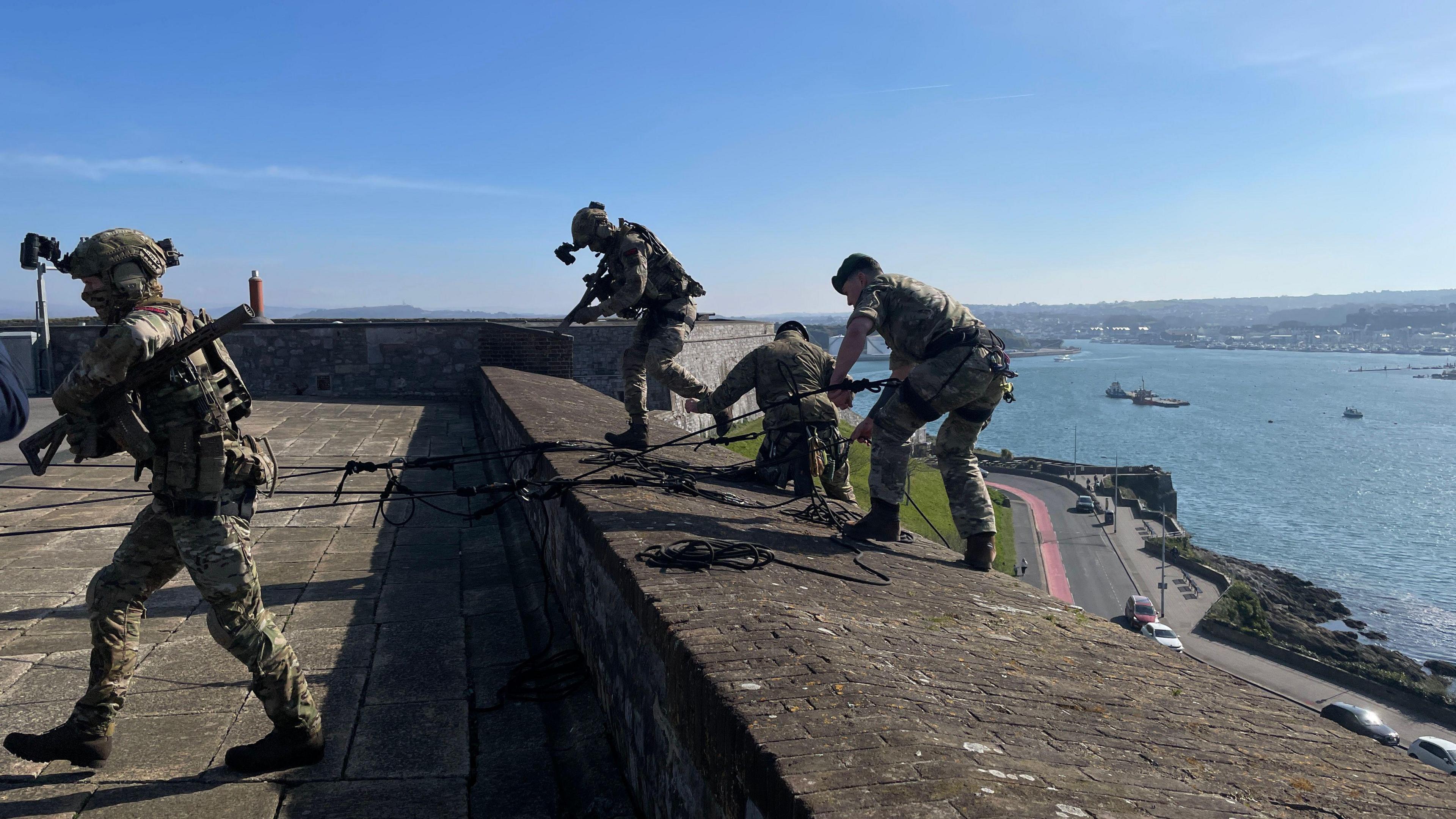 Military personnel on top of the Citadel in Plymouth. Three people are stood on the ledge abseiling and another walking to the left. Below is a road that curves around the Citadel and a harbour.