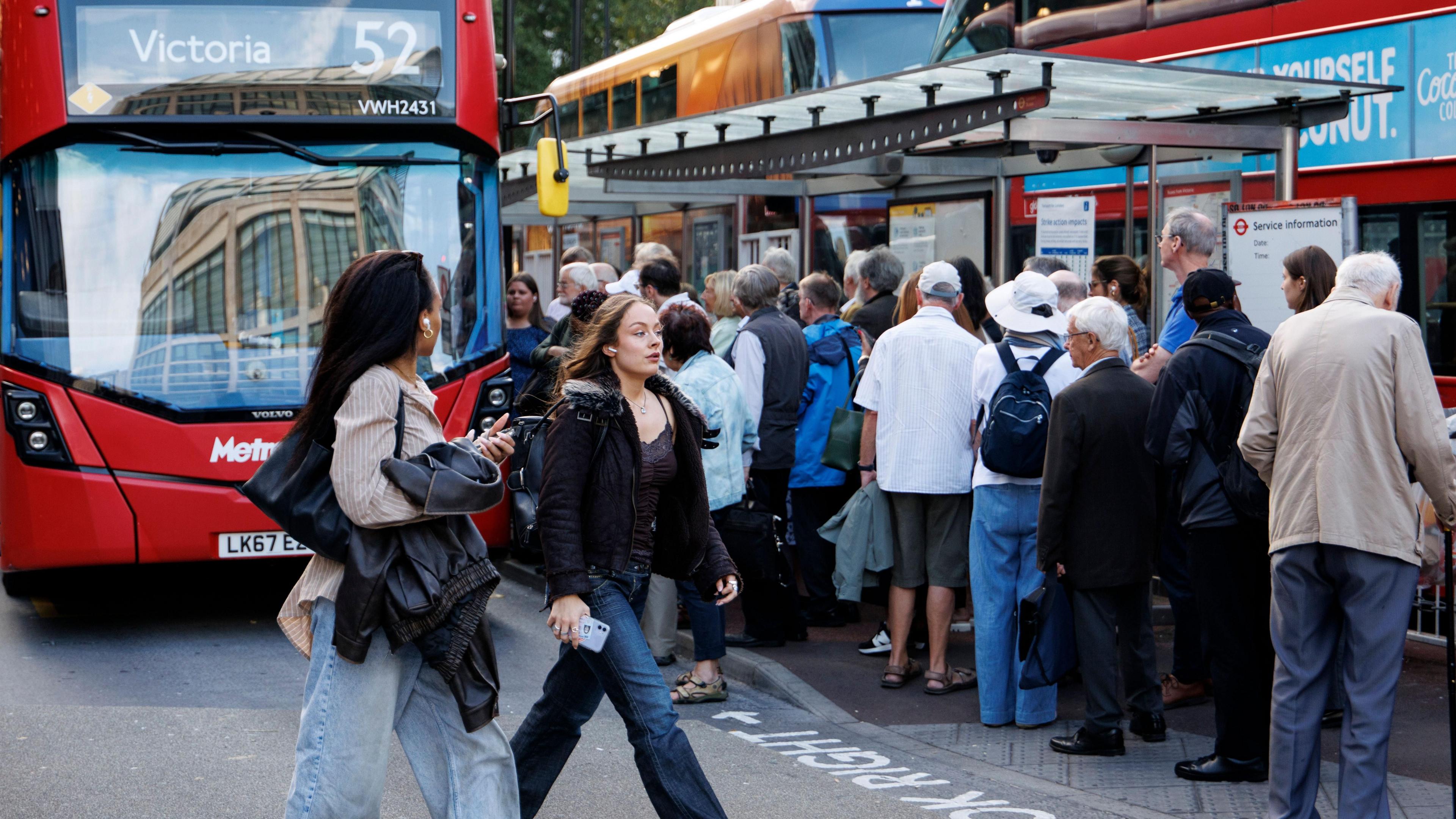 A large group of people queue at a busy London bus stop next to red double-decker buses, with two women walking past in the foreground.