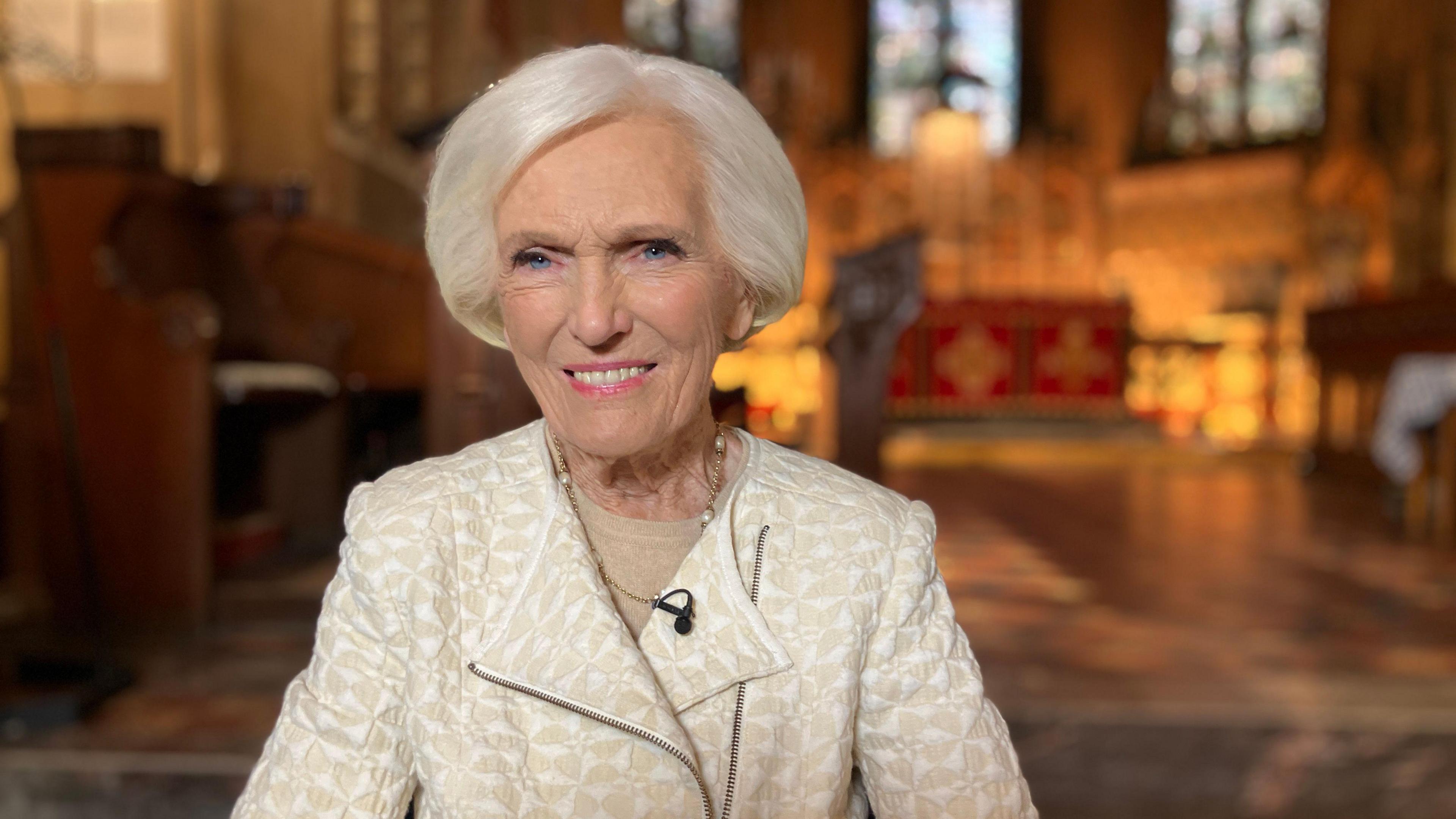 Mary Berry is sitting and smiling. She is wearing a beige and white jacket and a gold necklace. She has white hair in a short bob. 