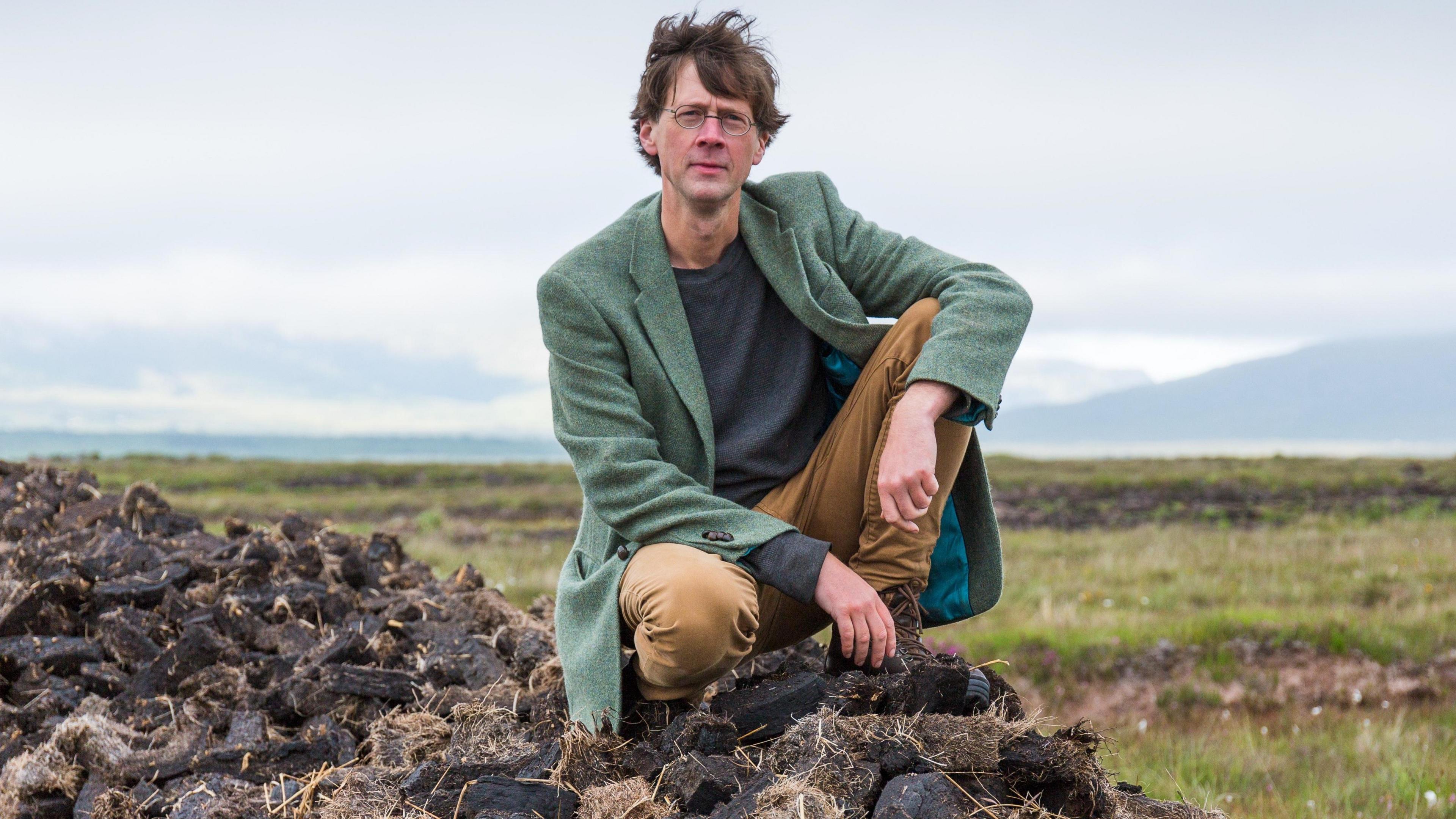 A man in a long green jacket, brown trousers and a blue jumper. He has brown hair. There are mountains behind him. He is kneeling down.