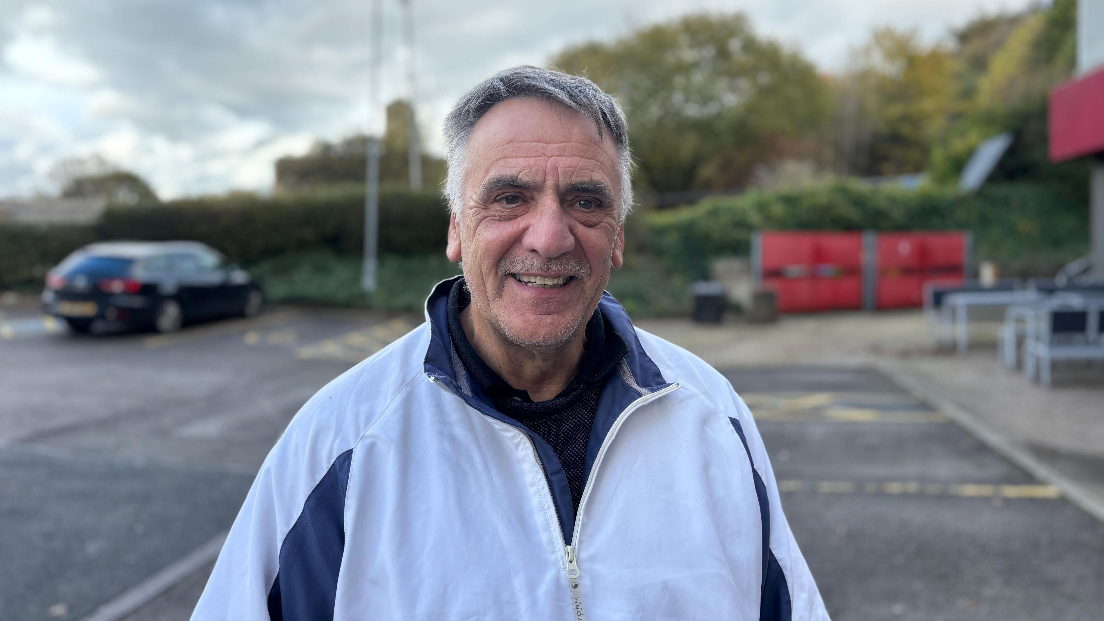 A man in his 60s wearing a white zipped tracksuit top. He is smiling directly at the camera.