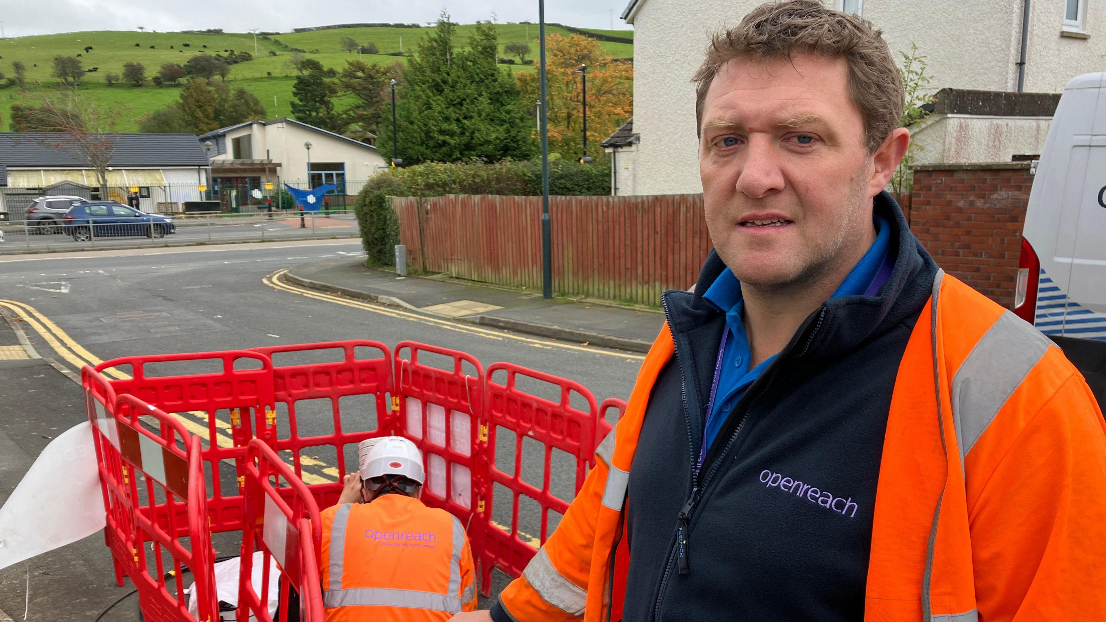 Tegryd Rees wearing an Openreach uniform and high-viz jacket, with a colleague doing work in the background