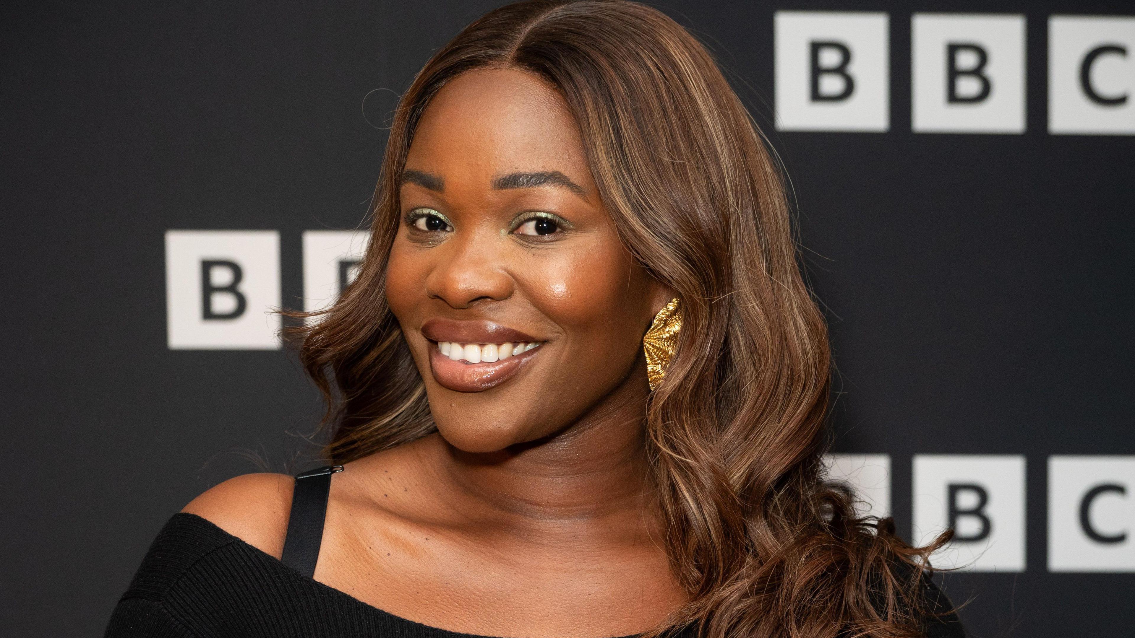 A woman with brown hair and a large gold earing smiles at the camera with the BBC logo on a background behind  her.