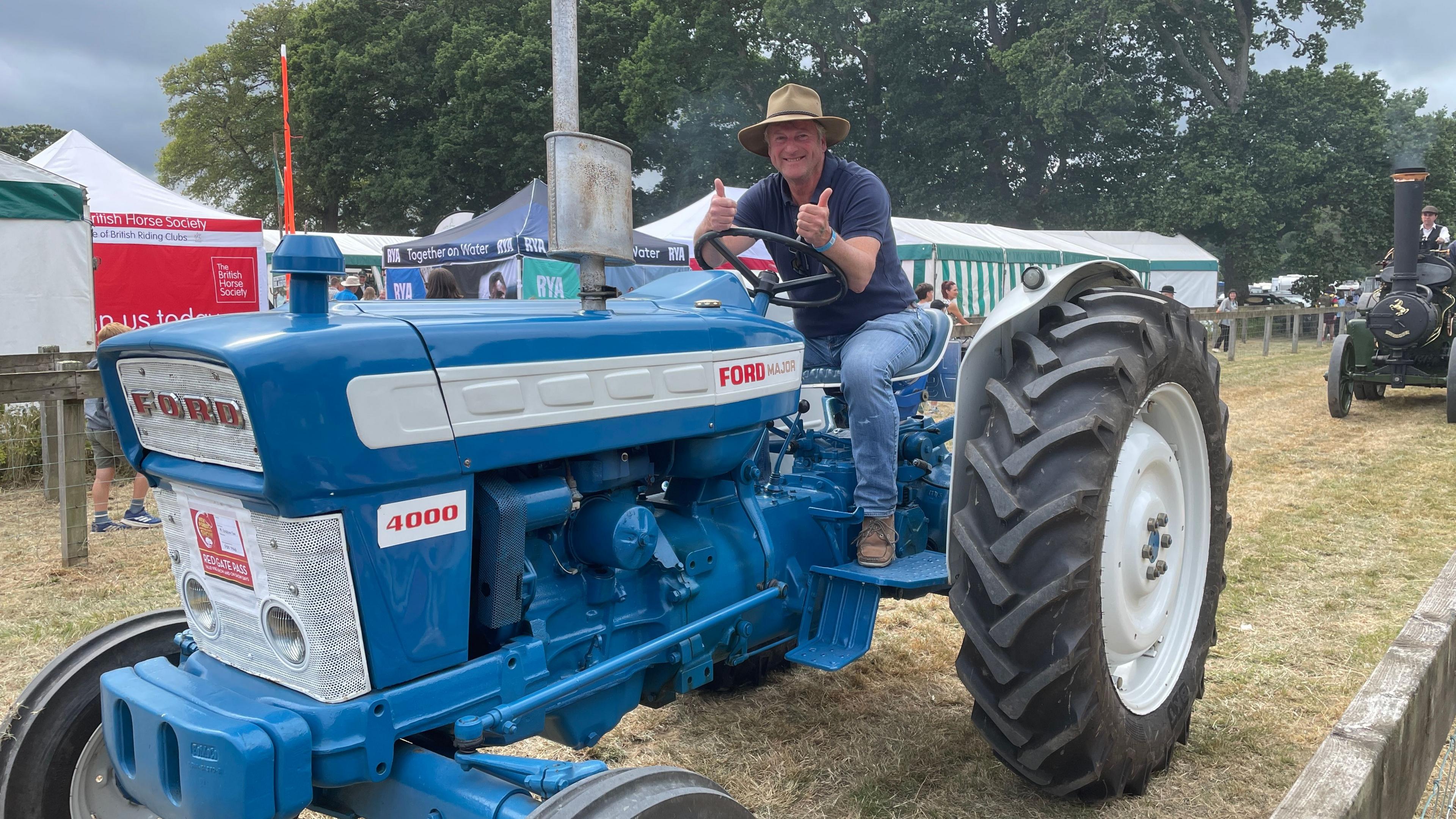 A man sits on a blue tractor with his thumbs up and smiling for the camera. 