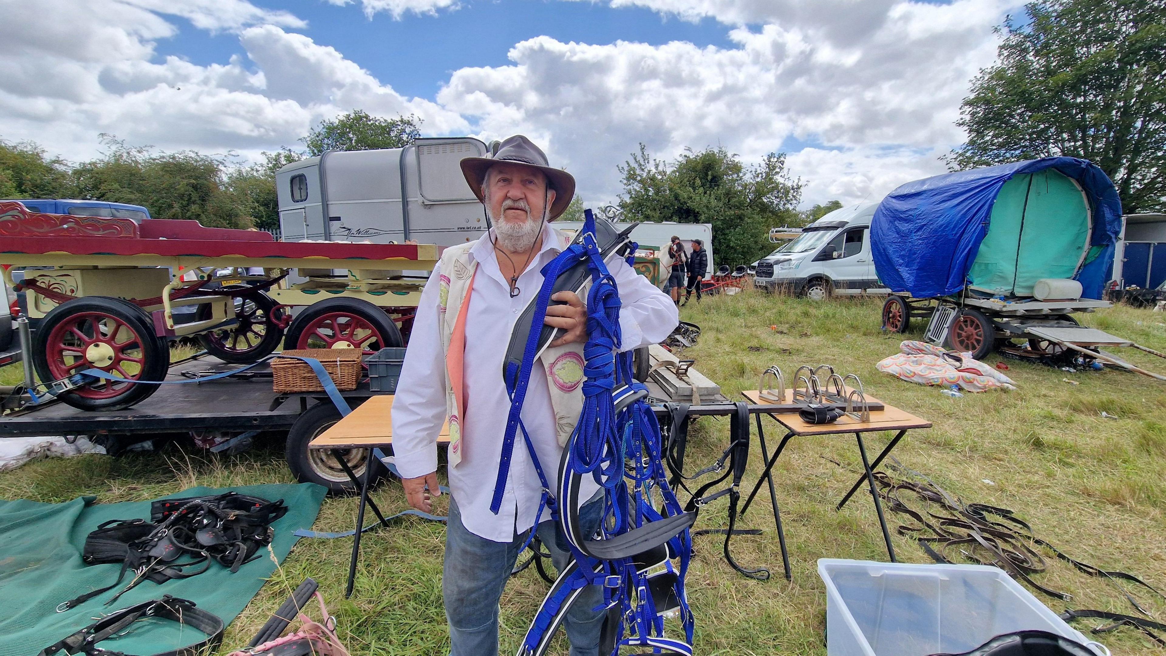 Man in a hat, waistcoat, shirt and jeans holding a blue harness. There are harnesses, carts, vehicles and people behind him on grass.