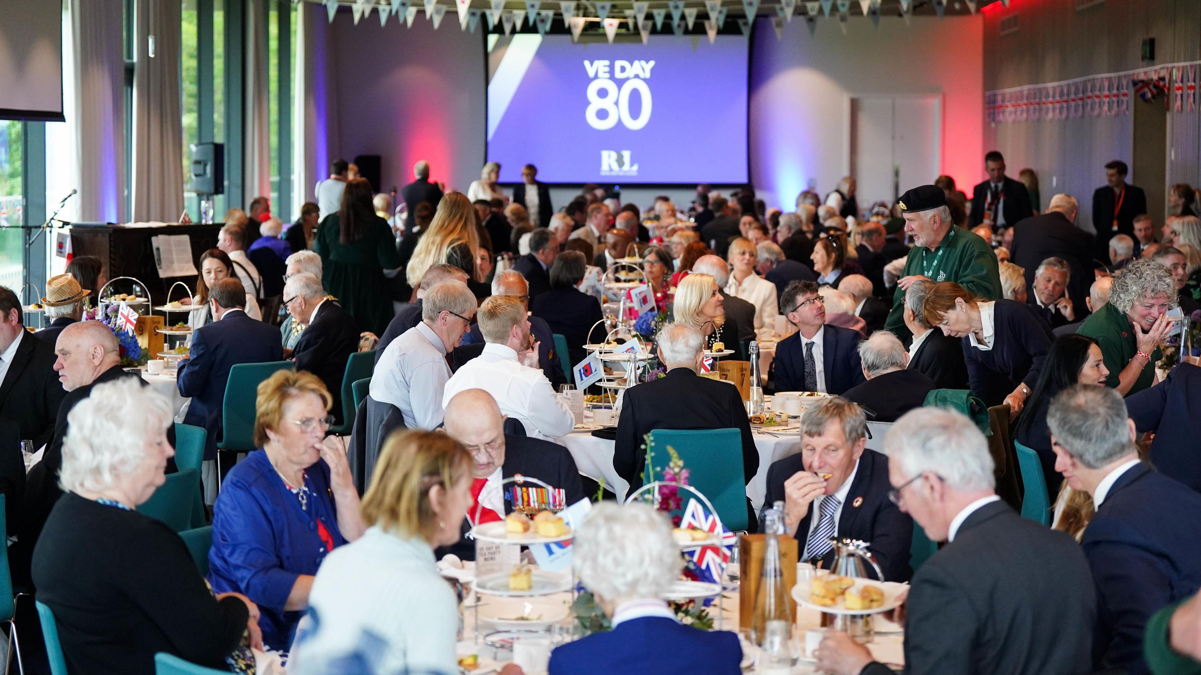 A room full of people sitting at tables during a tea party. A screen on the far wall says "VE Day 80".