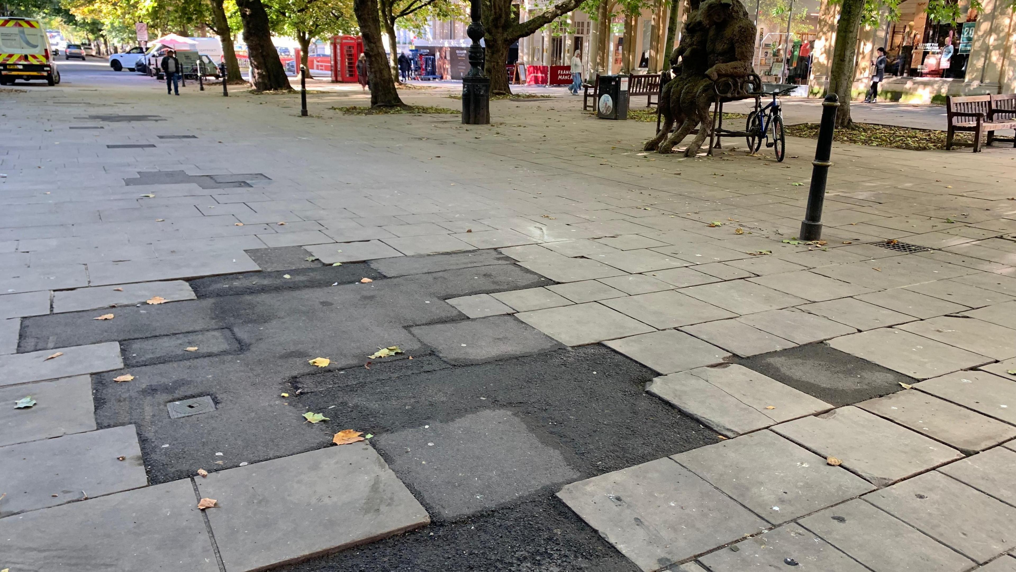 A close-up of the 'tarmac cowpat' on Cheltenham's Promenade. A large patch of asphalt sticks out, surrounded by stone paving slabs. Shoppers can be seen walking in the distance. There are trees lining the Promenade, as well as benches and statues.