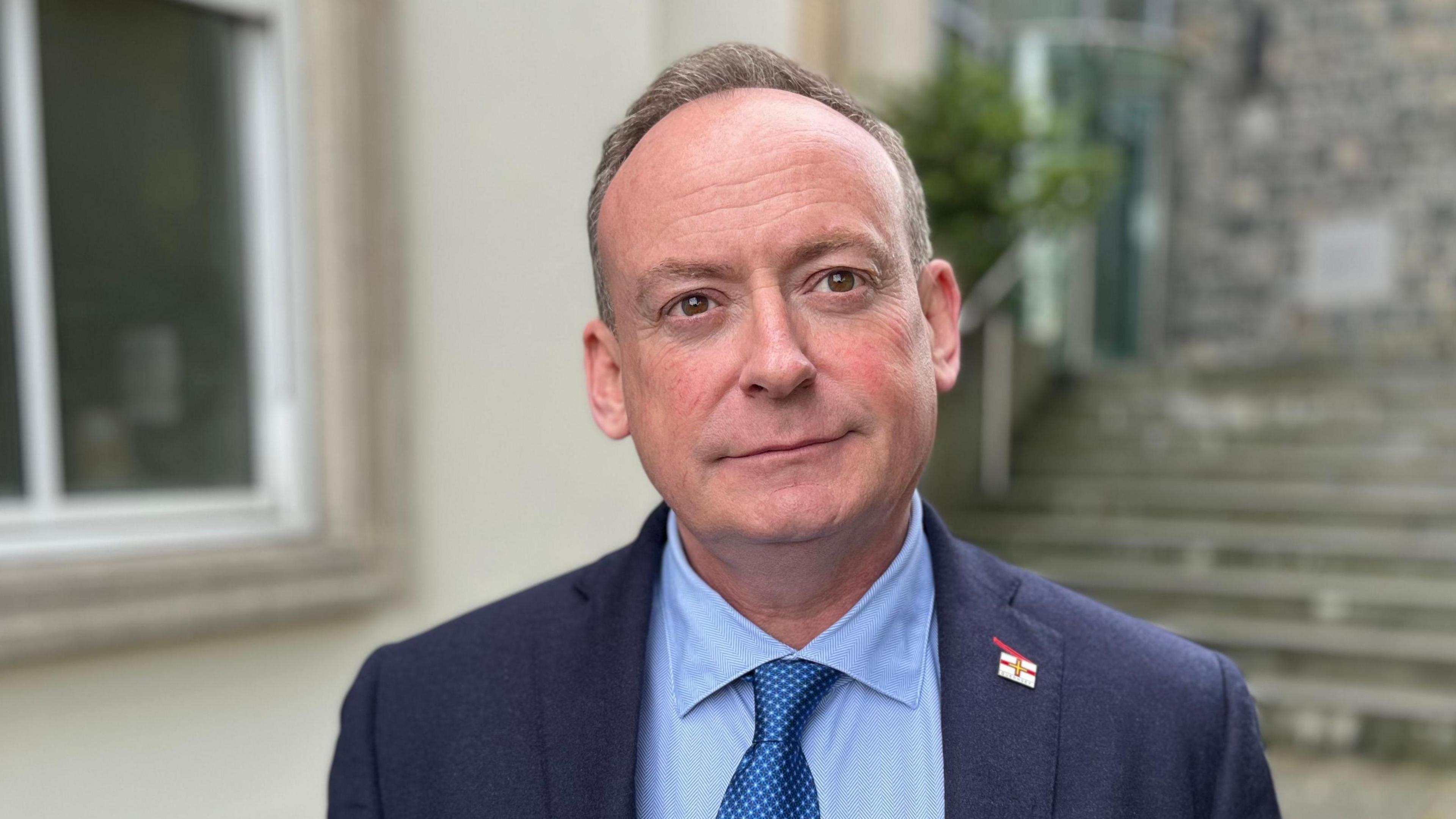 A man with short brown and grey hair looking towards the camera. He's wearing a blue suit jacket, with a blue shirt and a metallic blue tie. On the lapel of his suit jacket is a Guernsey flag pin badge.