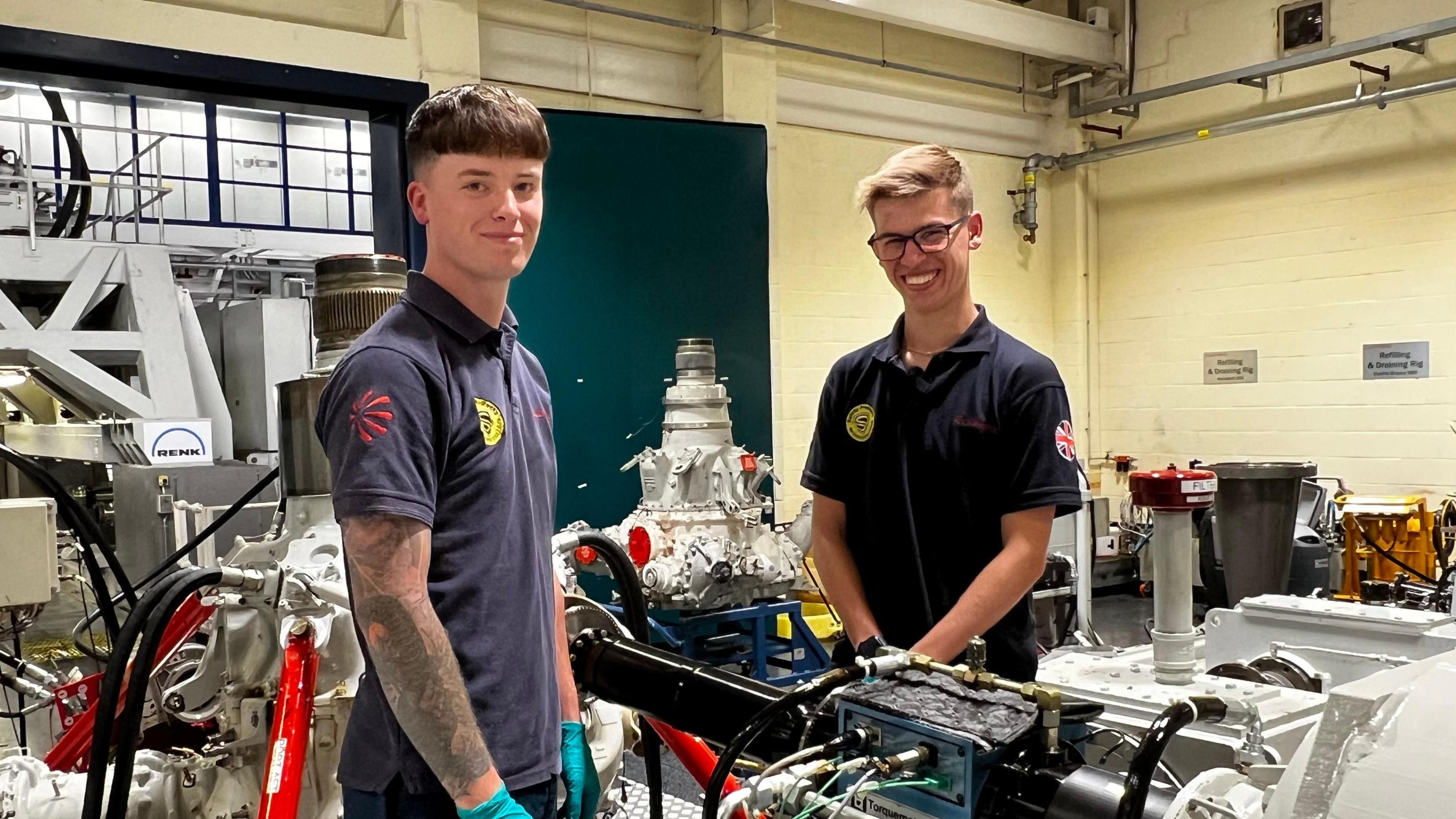 Two men in a factory smile at camera. There are various engine parts behind them. Both are wearing blue tops, and are younger workers. The factory walls are painted yellow.