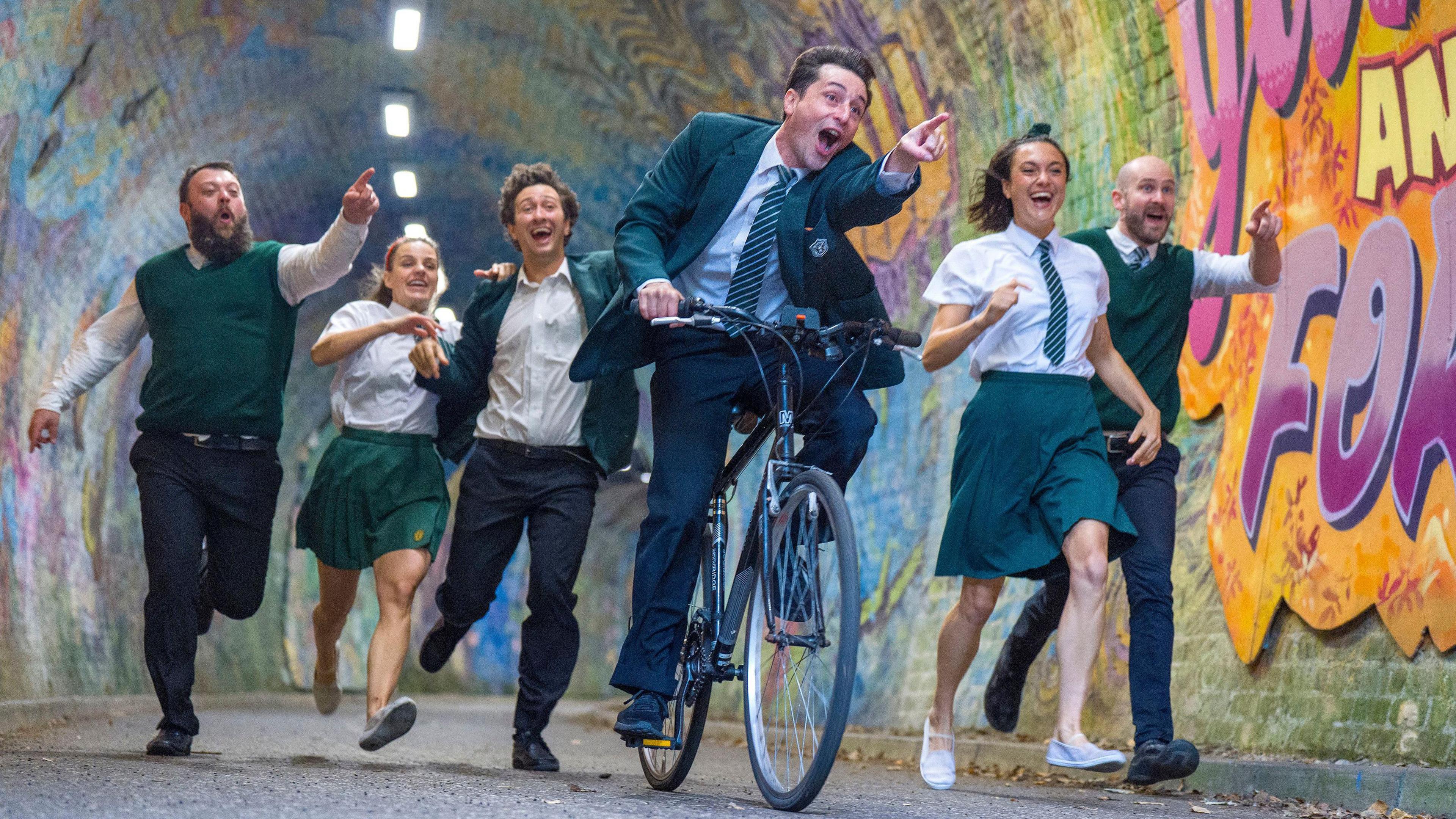 Six members of the Theatre Re ensemble cycle ad run through the Colinton Tunnel in Edinburgh. A young man in a green school uniform leads the way on a bike, pointing ahead and two girls and three men run alongside him, smiling and pointing.
