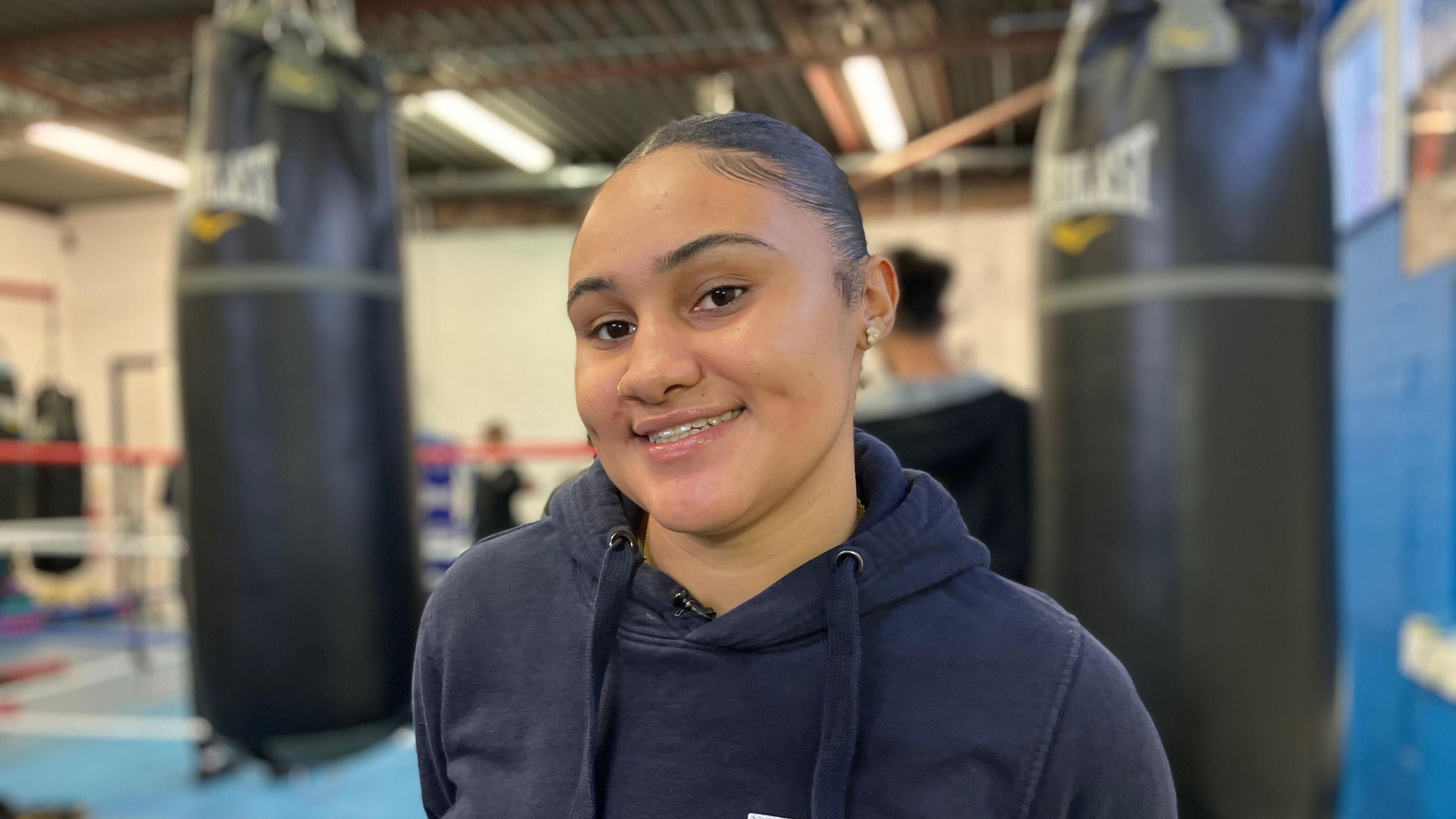 Serena Wiebe is standing in a gym. She is wearing a blue hoodie and is smiling at the camera. Behind her is a boxing ring and large black punch bags are hanging from the ceiling.