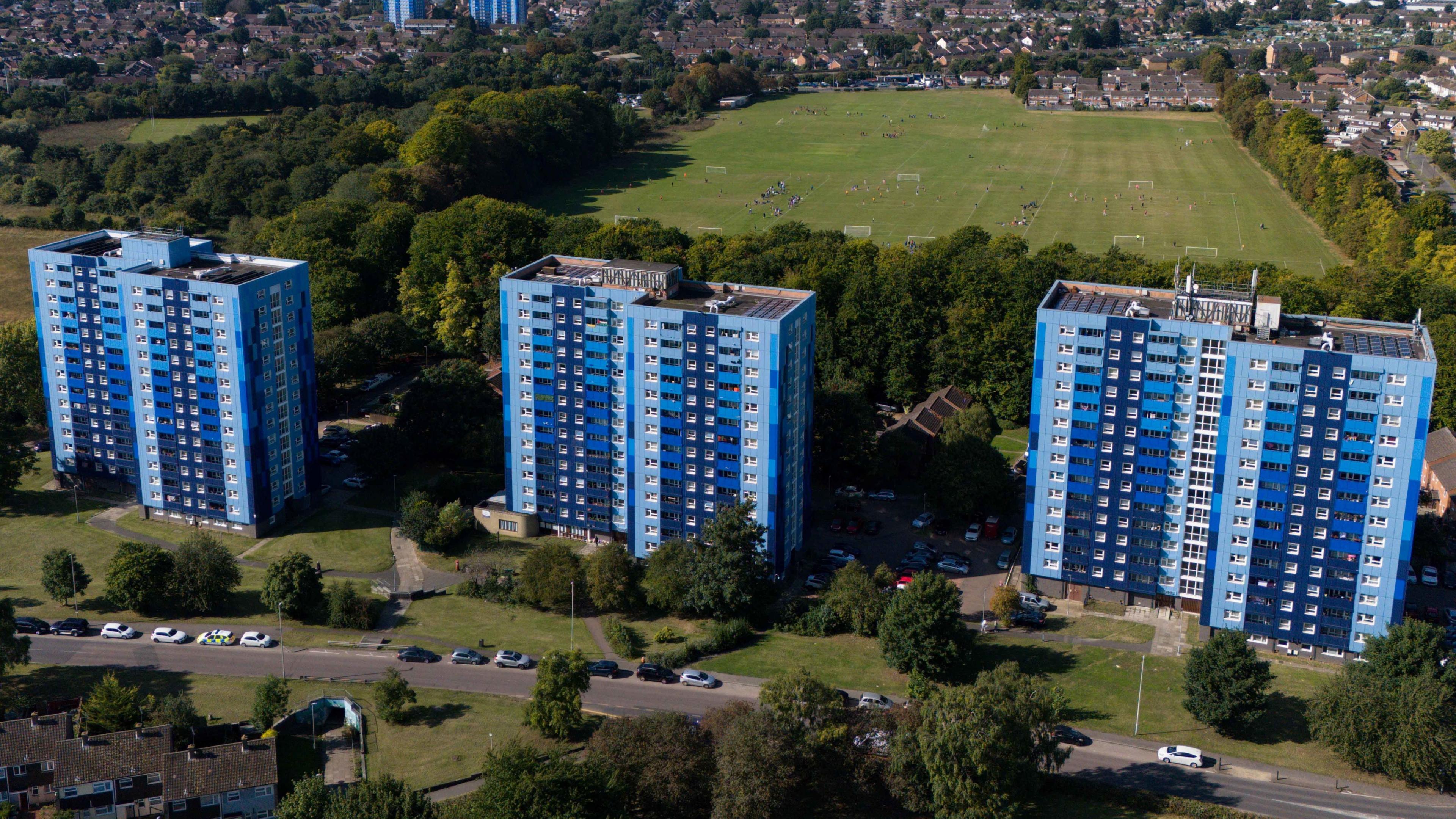 Three blue tower blocks surrounded by trees and grassy areas