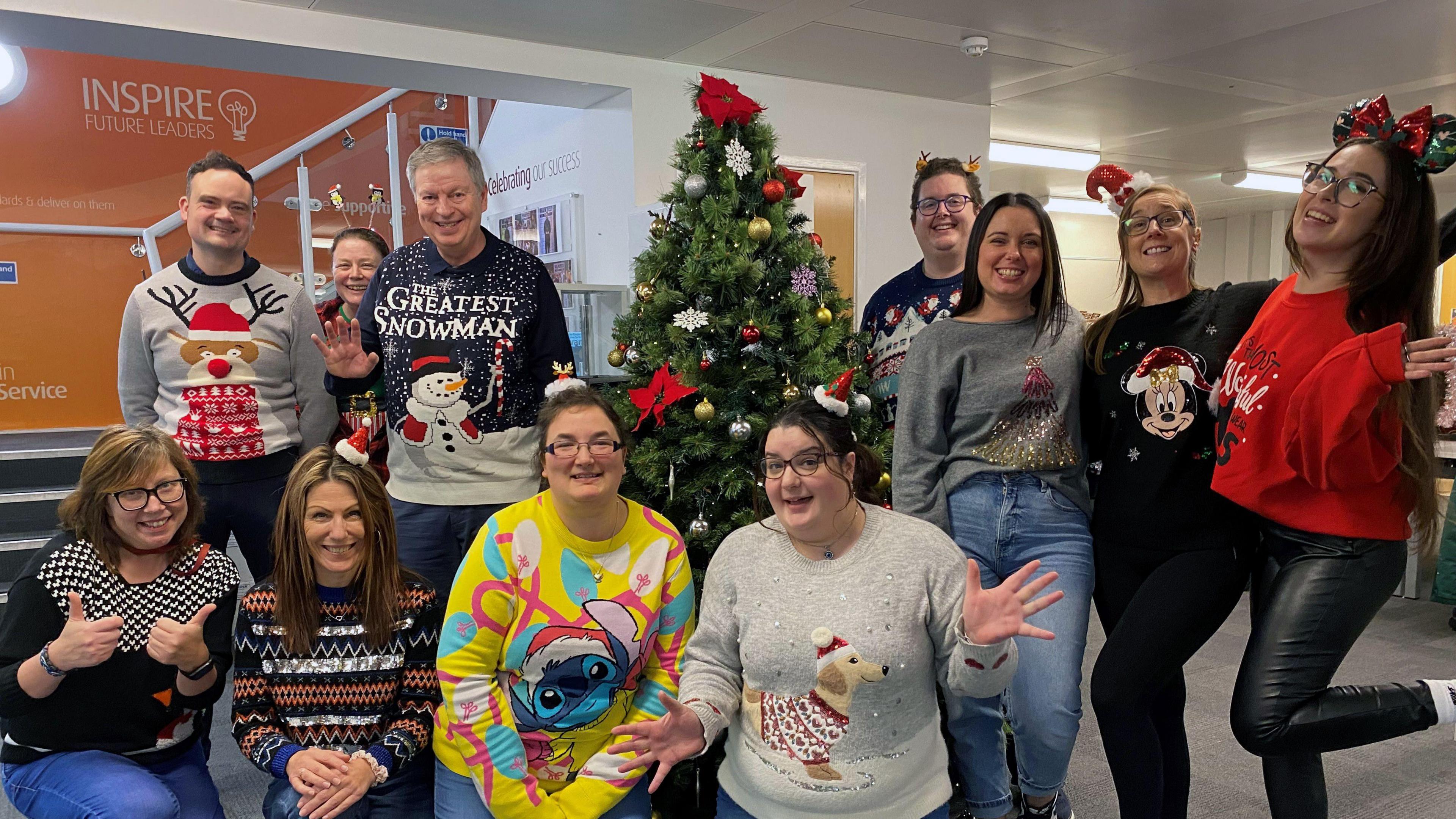 A group of workers from the UK Power Networks call centre in Ipswich are gathered together around a Christmas tree smiling. They wear Christmas hats, jumpers and other festive decorations on them.