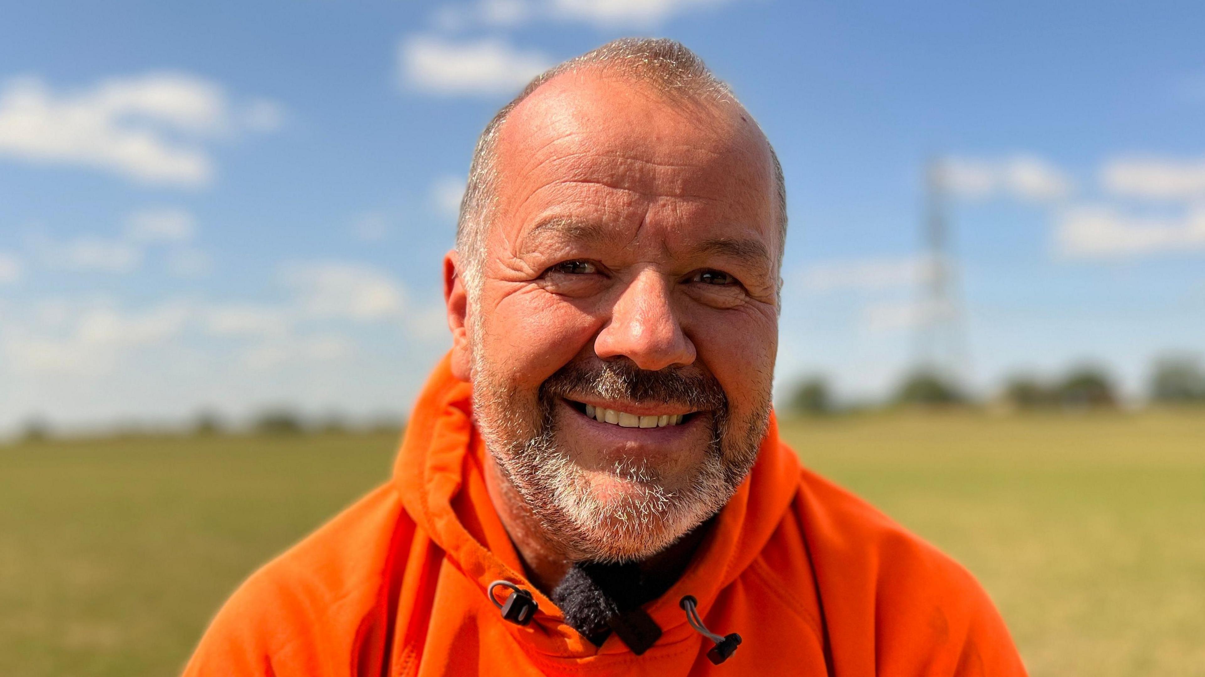 Richard Owens is wearing a bright orange hoodie. He is smiling. The field behind Richard Owens green with patches of yellow grass.