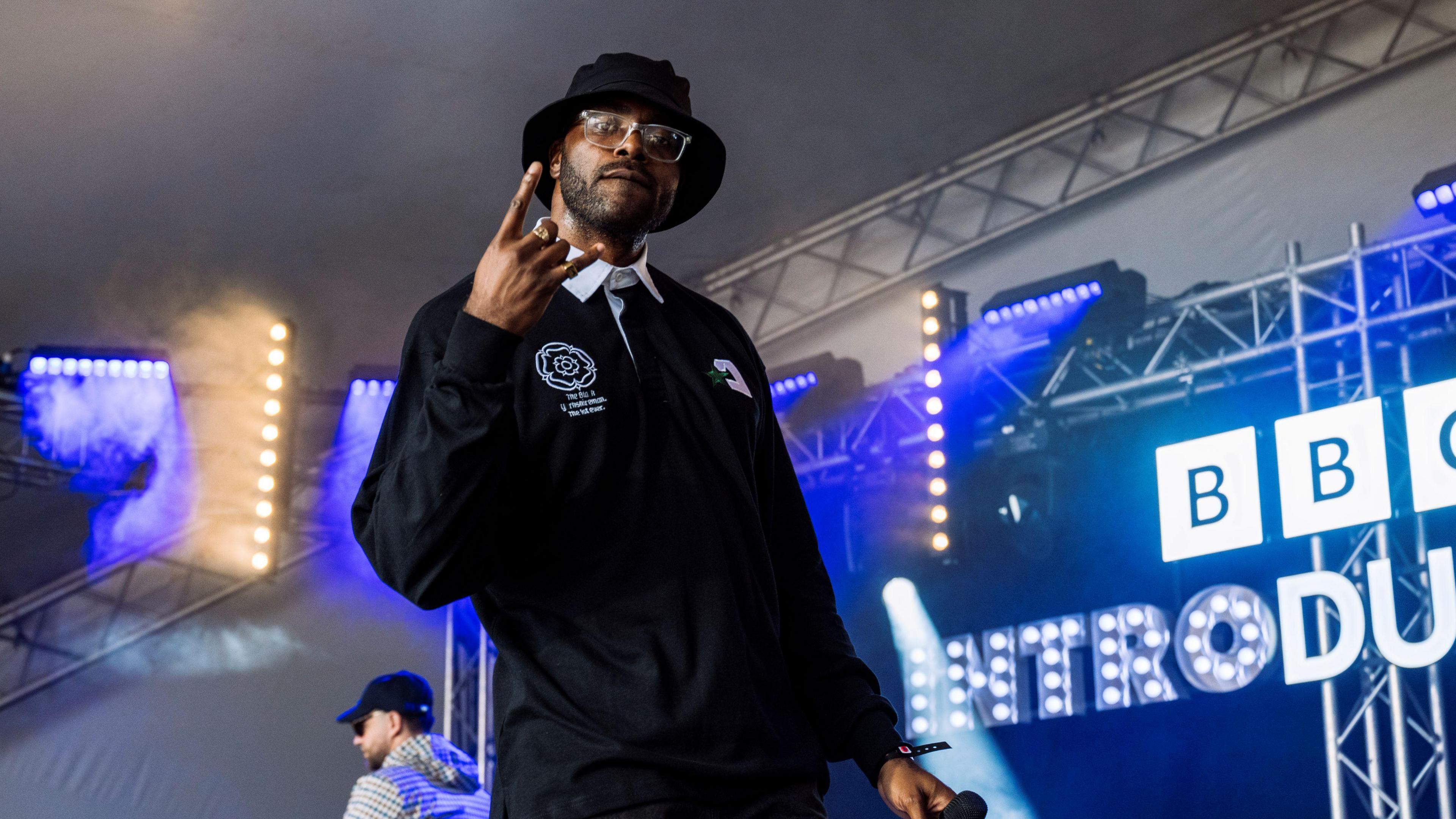 A man wearing a black bucket hat, clear-frame glasses, and a black long-sleeve polo shirt with a white logo and a white collar. He is standing on a stage with blue and white stage lights in the background and a white lit-up sign reading "BBC INTRODUCING" in capital letters.