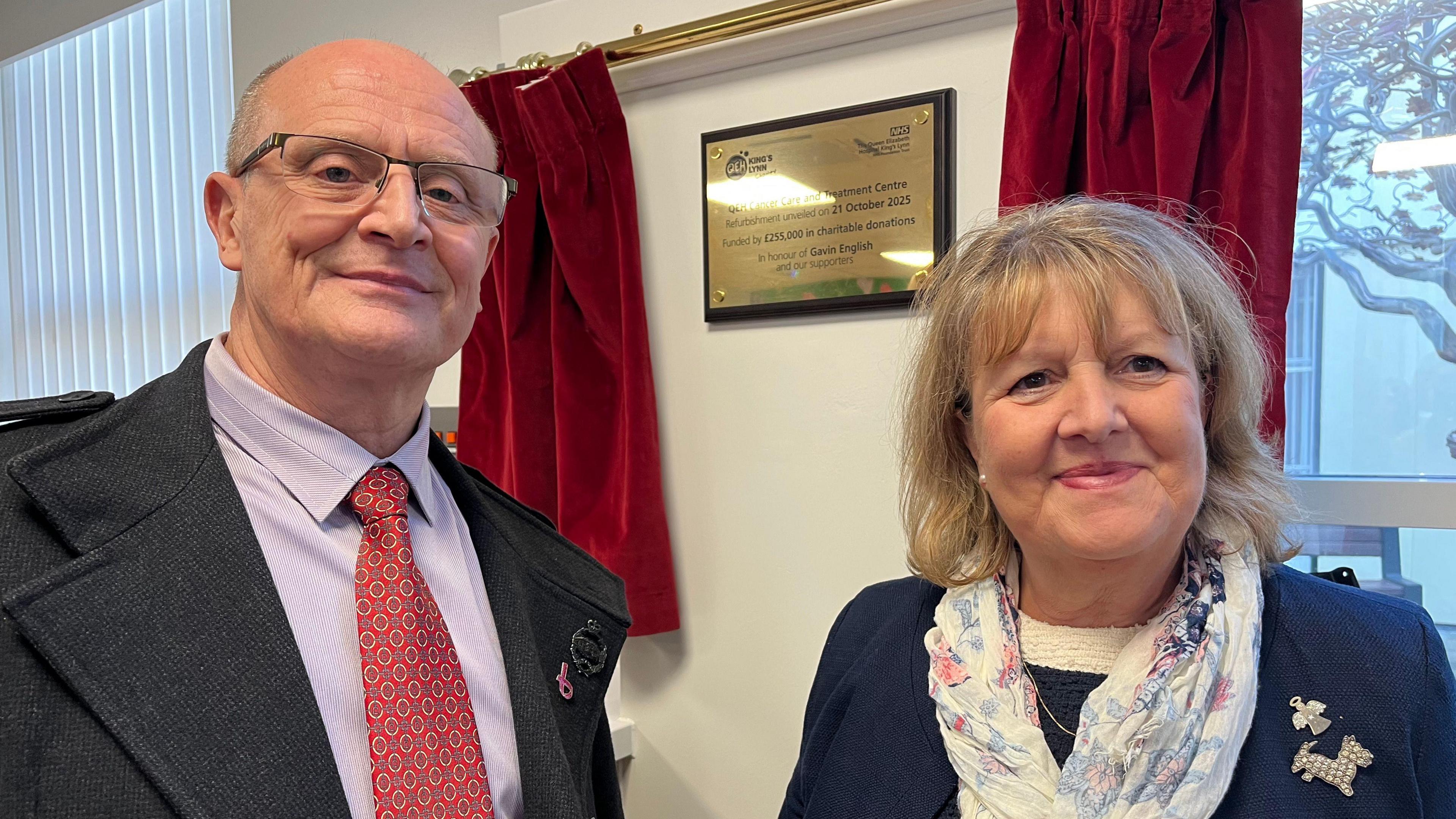 Belinda Penkethman and Ray English stand in front of a shiny brass plaque commemorating their father Gary English. Ray is wearing a charcoal coat, pink shirt and red tie. Belinda wears a navy top and a white patterned scarf.