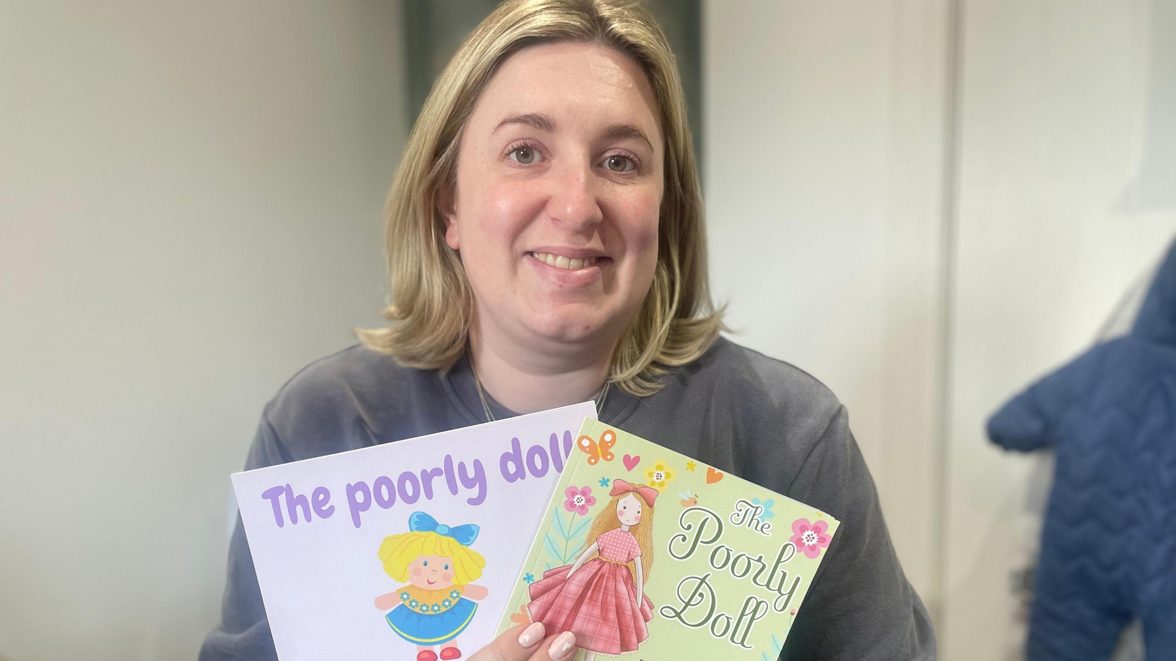 Lauren Bolus-Jones smiling while holding up to versions of a book called The Poorly Doll. 