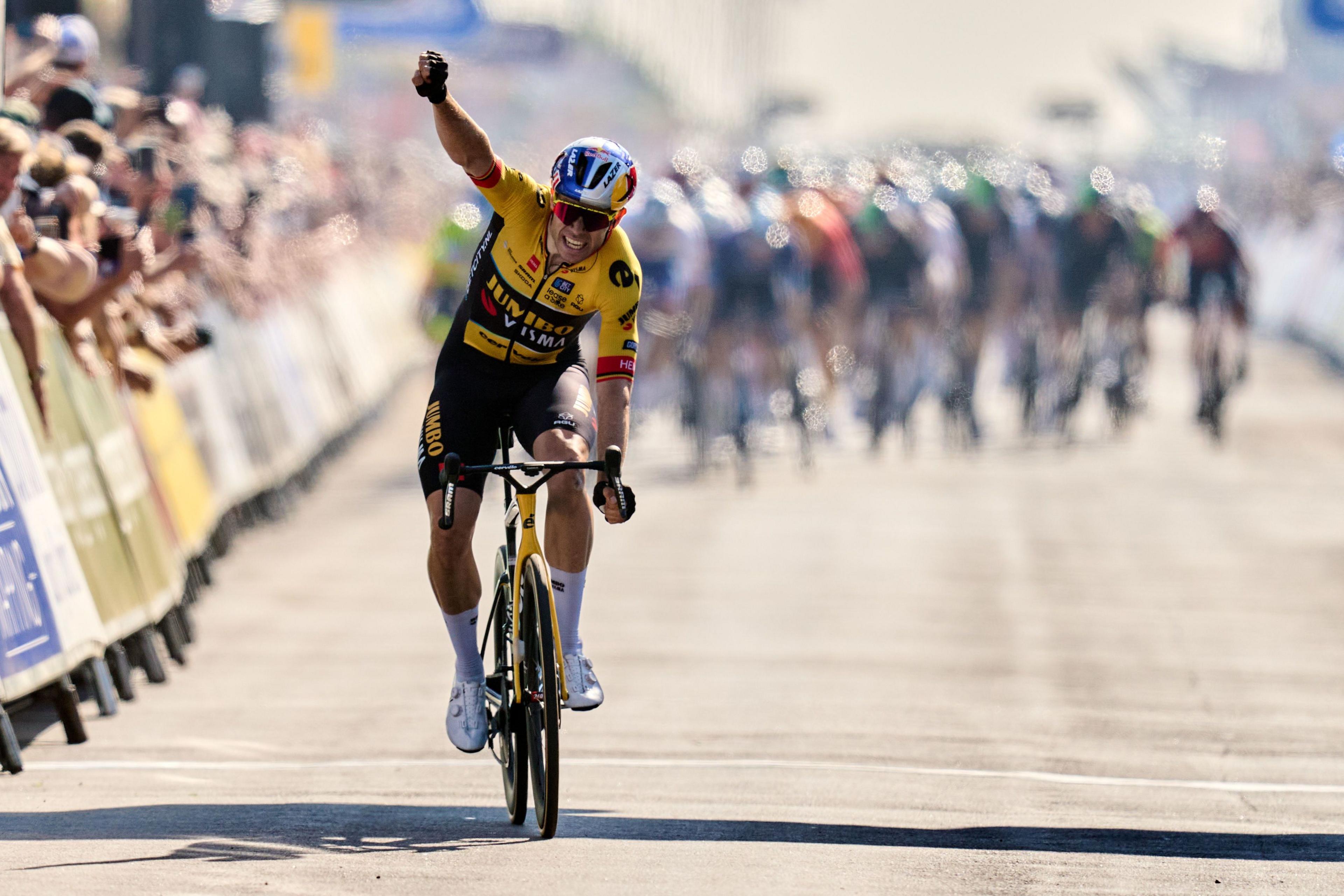 Cyclist Wout van Aert is pictured crossing the final stage finish line of the Tour of Britain in 2023, which he won. He is wearing a yellow and black cycling suit while riding his yellow bike.