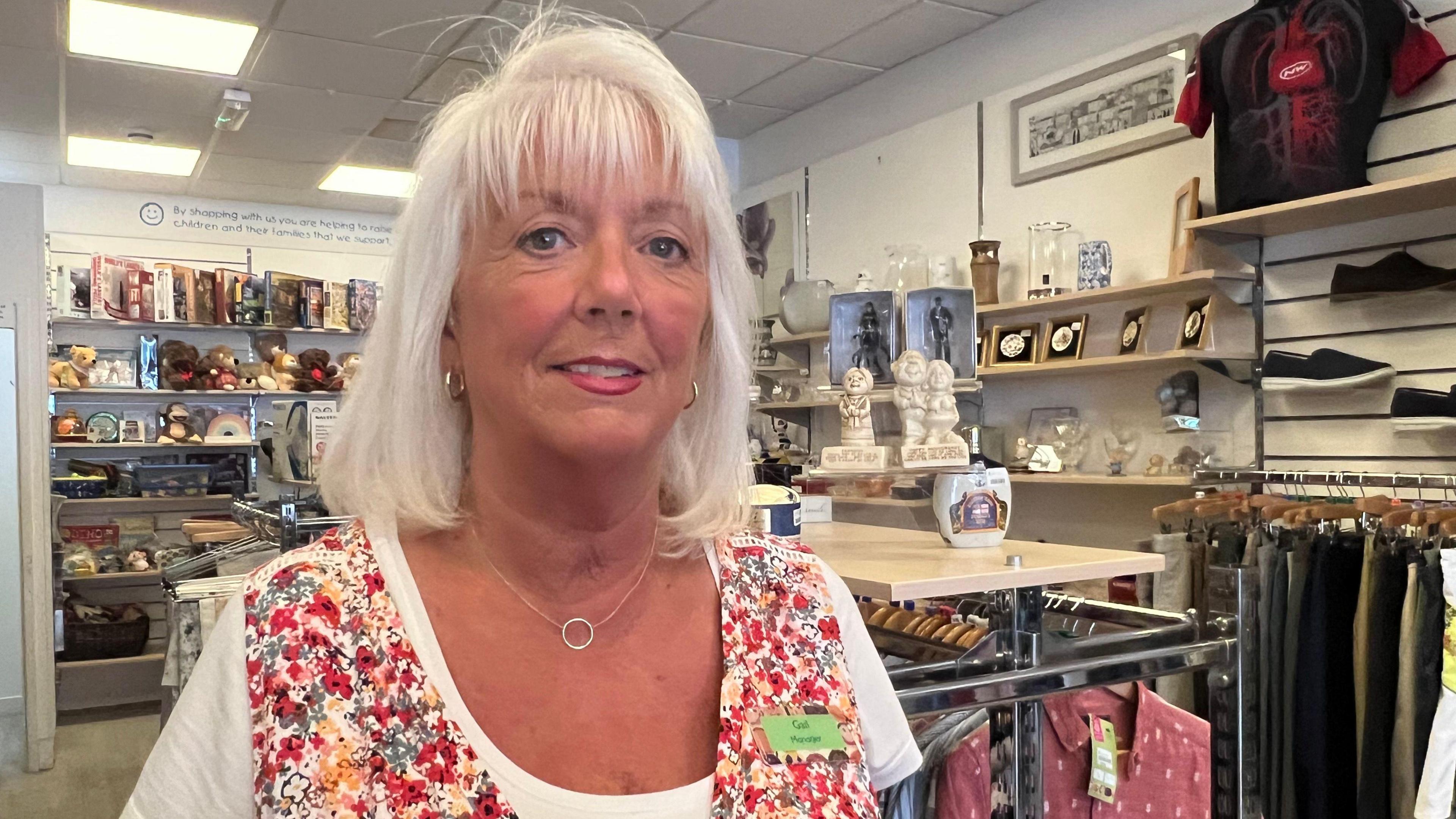 Gail Fry inside a charity shop. She wears a white T-shirt and a floral vest, and has white hair cut into a bob with a fringe. 