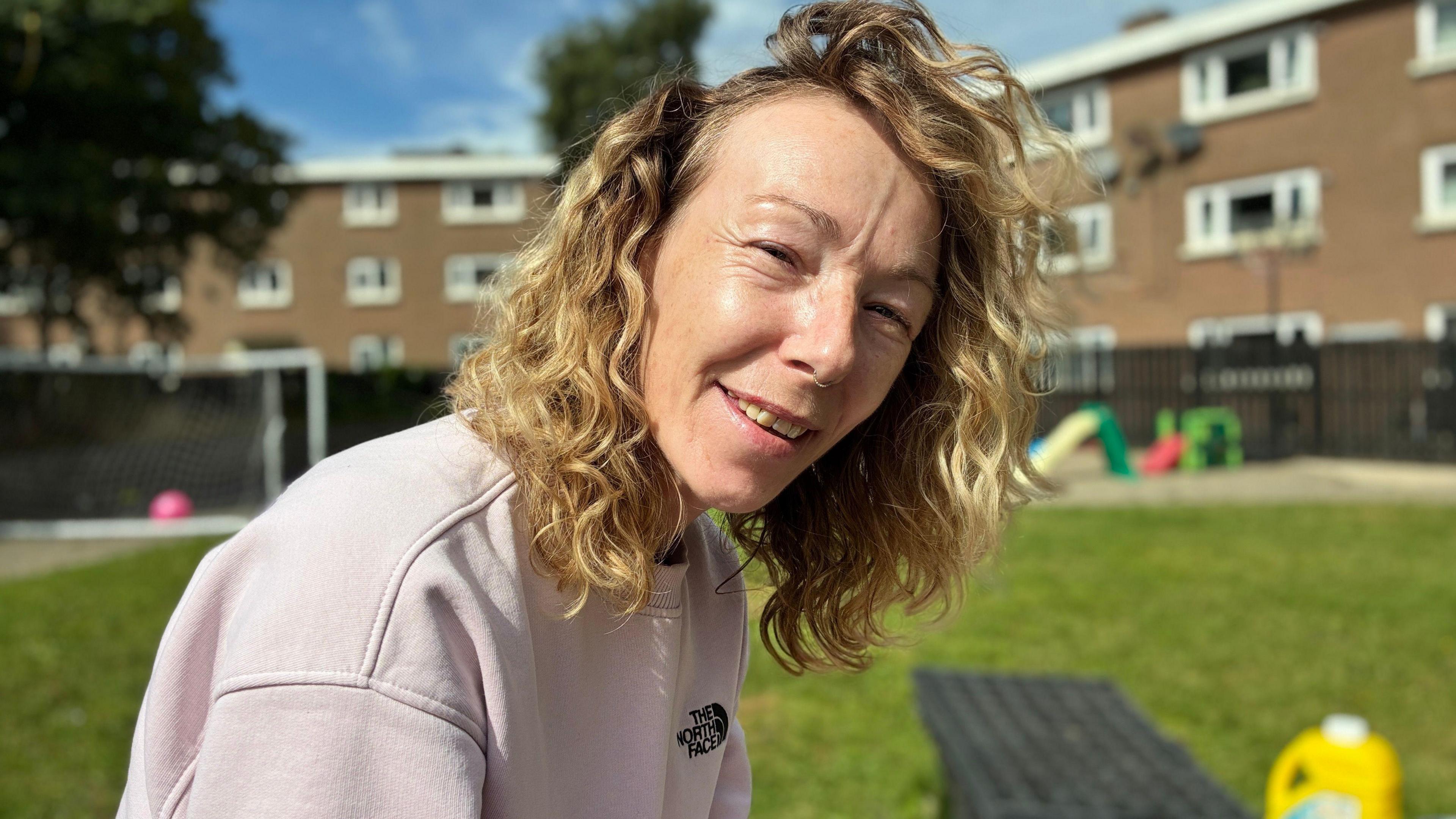 A woman with curly blonde hair smiles outdoors on a sunny day. She’s wearing a pale pink sweatshirt with a visible “The North Face” logo. Behind her, a grassy area features a football goal, colourful playground equipment, and residential buildings with white-framed windows. The bright light and relaxed pose create a friendly, community atmosphere.